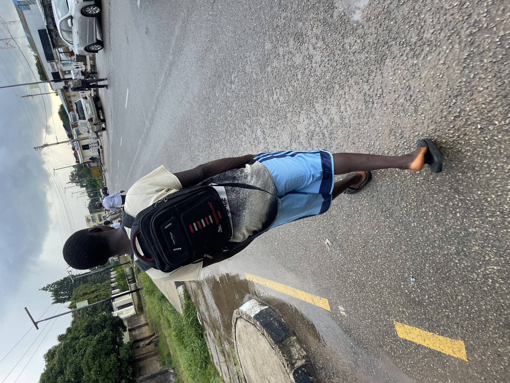 The image shows a young Black male from behind, walking on a wet, asphalt road in Jos, Nigeria. He is wearing a light-colored t-shirt, blue shorts with dark stripes, and dark sandals. A black backpack with red accents is slung over his shoulders. The road appears to be a main thoroughfare with painted yellow lines marking lanes. To the left of the road, there is a sidewalk with overgrown grass and a concrete curb, with a puddle of water on the road surface. In the background, there are buildings, streetlights, and several vehicles, including a silver sedan and a light-colored SUV, as well as a motorcycle. The sky is overcast with clouds. The overall impression is a typical street scene in Jos, likely after a rain shower.