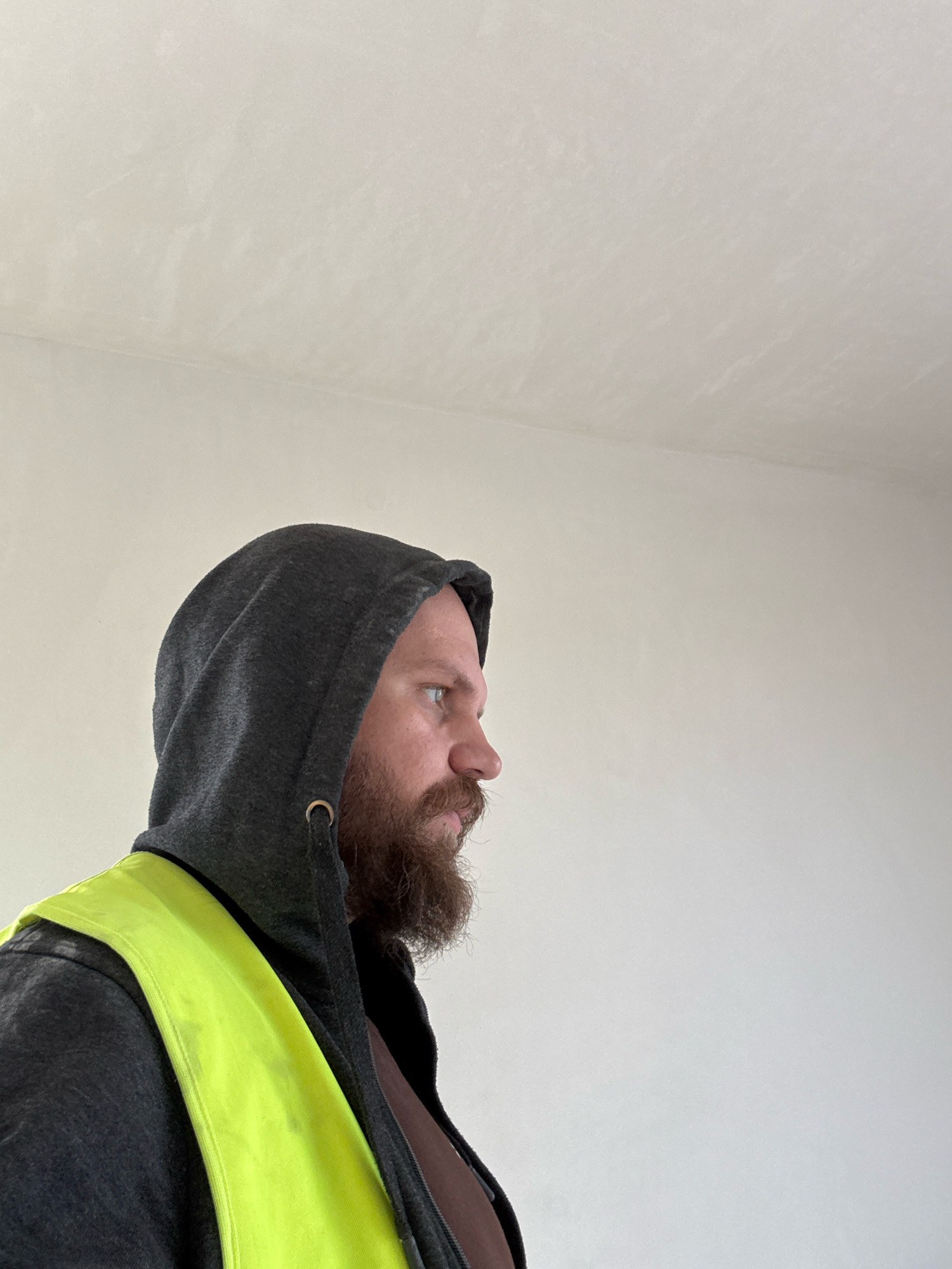 A medium shot shows a man in profile, facing right, wearing a dark gray hooded sweatshirt and a bright yellow safety vest. He has a full beard and a mustache. The background is a plain, light-colored wall and ceiling. The image appears to be taken indoors in what could be a construction or renovation site, given the safety vest. There are no indications of the specific location in Nowa Sól, Poland.