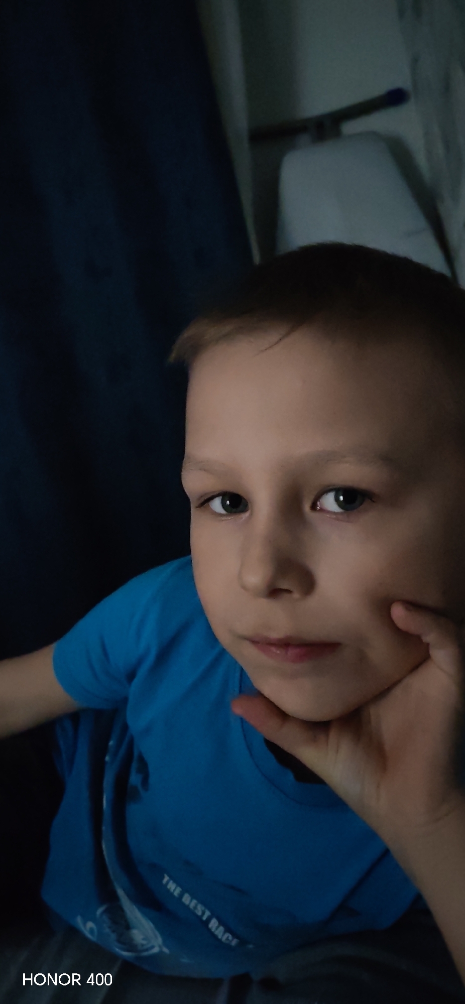 A close-up shot shows a young boy with short, light brown hair and light eyes. He is wearing a blue t-shirt with white writing on it that reads "THE BEST RACE". His left hand is supporting his chin. The background includes a dark blue curtain and what appears to be a white object with a dark rod attached. The photo was taken with an HONOR 400 phone. There is no information to suggest the location is Noviy, Russia.