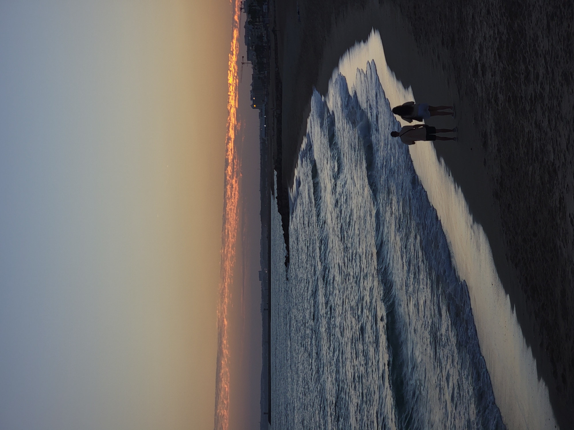Two individuals, silhouetted against a twilight sky, walk along the sandy shore of Quarteira, Portugal. The ocean, a deep blue, gently laps the coast, leaving a reflective sheen on the wet sand. In the distance, the urban landscape of Quarteira emerges from the fading light, with buildings and a long pier punctuating the horizon. The sky above is a gradient of soft blues and oranges, with wisps of clouds catching the last rays of the sun. The scene captures a serene moment of a couple enjoying the tranquil beauty of the Portuguese coastline at dusk.