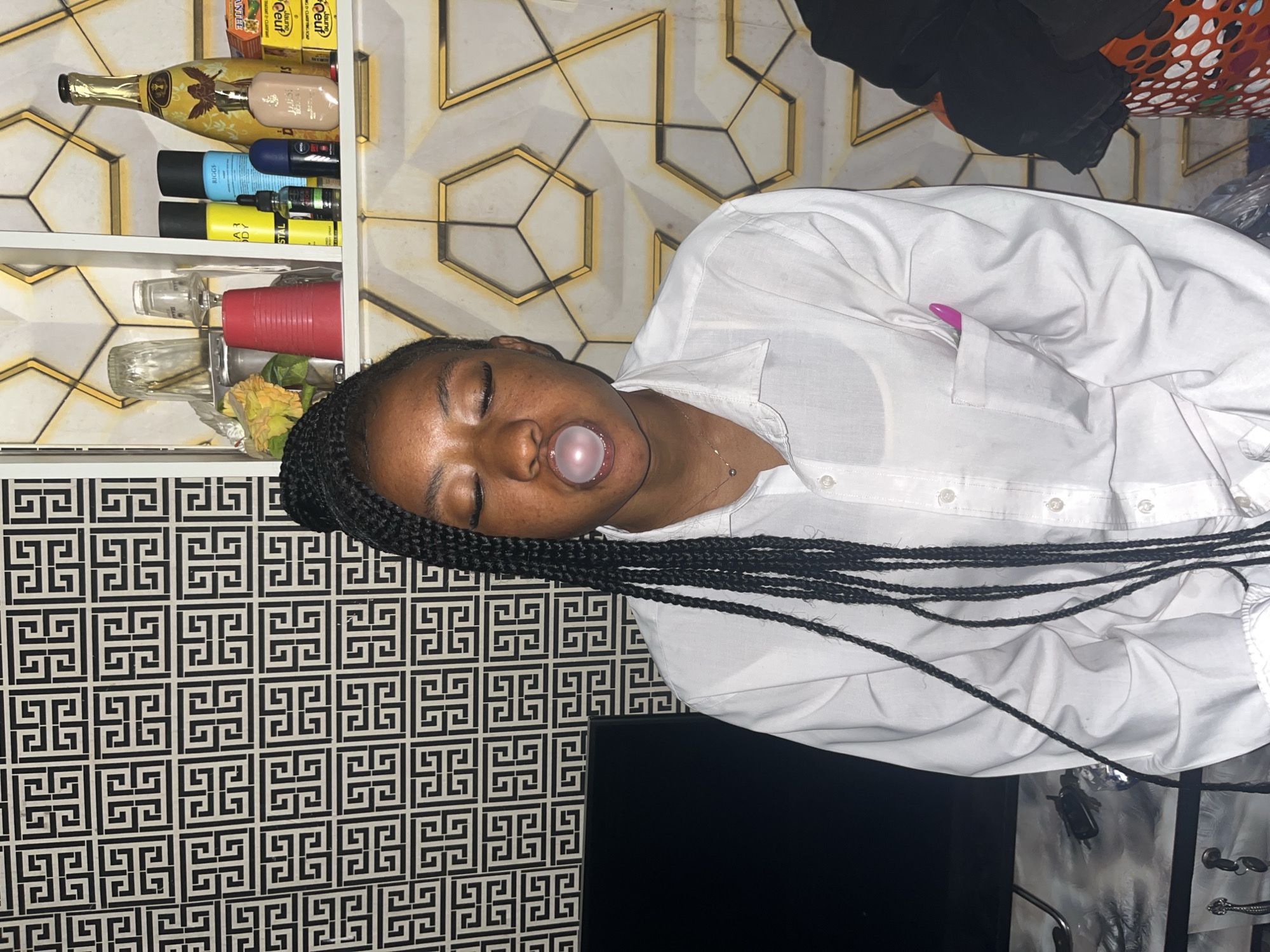 The image shows a young woman with dark skin and braided hair, blowing a bubblegum bubble. She is wearing a white button-down shirt. Her eyes are closed and her head is tilted back. The background features a shelf with various items, including bottles of liquid, and a patterned wallpaper. Below the shelf is a black and white patterned wall. A black television is visible at the bottom of the frame. The overall scene appears to be indoors, likely in a home or a store. The lighting suggests it could be daytime or an indoor setting with artificial light.