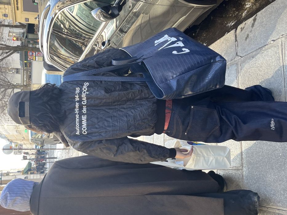 The scene depicts two individuals walking on a stone-paved sidewalk in Paris, France, likely on or near Rue de Monceau, indicated by a blurred street sign in the distant background. The foreground shows the back of a person with dark, wavy hair, wearing a reversed dark baseball cap. They are dressed in a black quilted bomber jacket featuring white text that reads "Automne-Hiver '84-'85 COMME des GARÇONS". A red patterned belt is visible above dark blue, wide-leg jeans with visible stitching and a small white embroidered logo on the lower left leg. This individual carries a large, dark blue, textured tote bag with "C.A.L.M" (or similar letters) in white block text on their left shoulder, and a plain white shopping bag in their right hand.
To the left, partially obscured, is another person seen from behind, wearing a long, dark coat and a light blue and white striped beanie or hood, also holding a white shopping bag.
The sidewalk appears slightly damp or reflective in areas. To the right, a dark grey SUV is parked or moving slowly, reflecting the surrounding architecture. In the middle ground, the street is lined with classic Parisian Haussmannian buildings, characterized by their light-colored stone facades, multiple windows, and occasional balconies. Various pedestrians and distant vehicles are visible. Street signs include a "BNP PARIBAS" sign on a building to the right and a "MONOPRIX" sign further down the street on the left. Traffic lights show green and red. The scene is illuminated by natural sunlight, casting visible shadows.