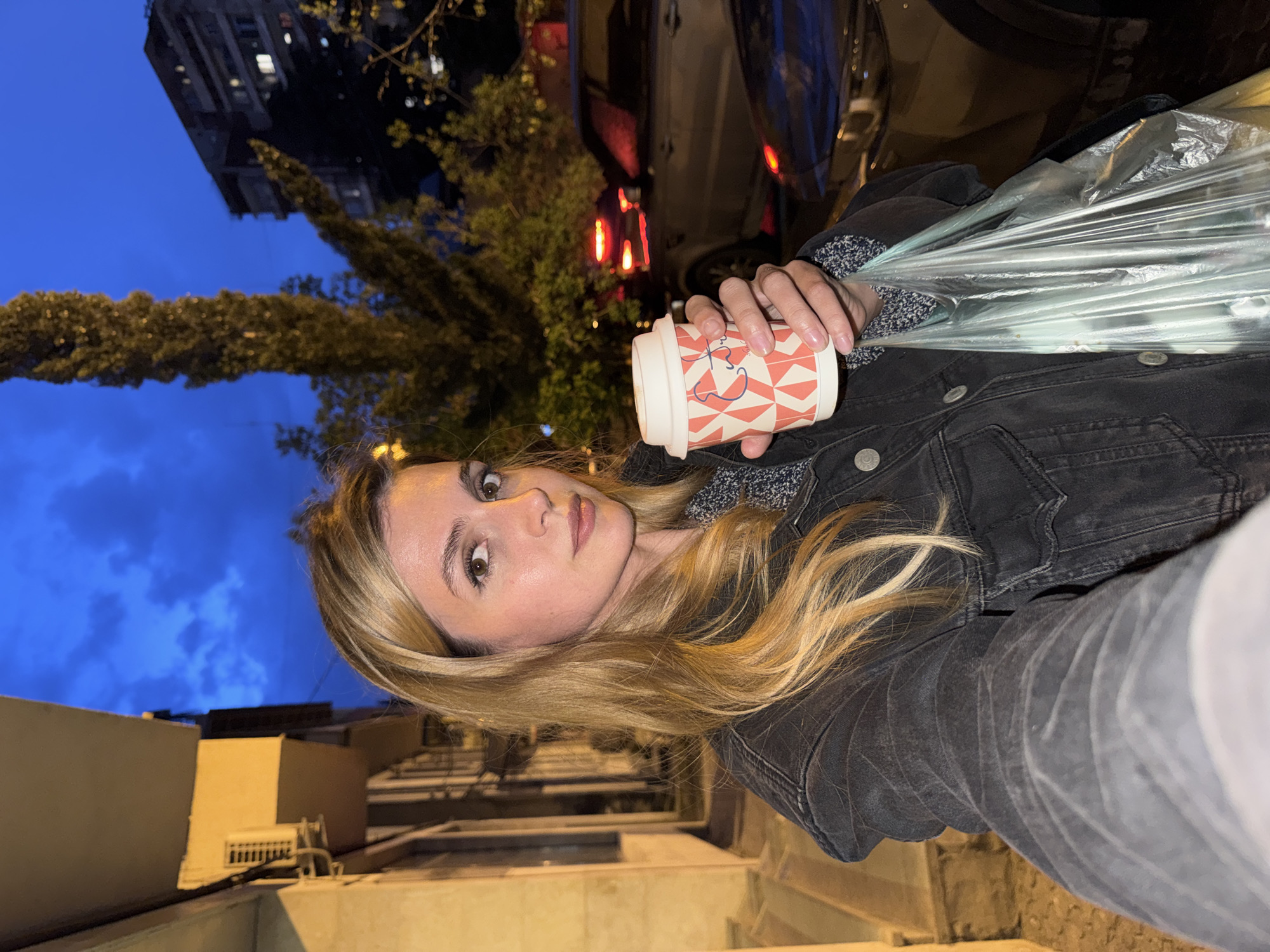 A woman is standing in Tbilisi, Georgia, holding a coffee cup and a plastic bag. She has blonde hair, and is wearing a black denim jacket. Behind her is a building, a tree, and a car. The sky is a dusk blue. The photo appears to be taken at ground level, outside on a street.