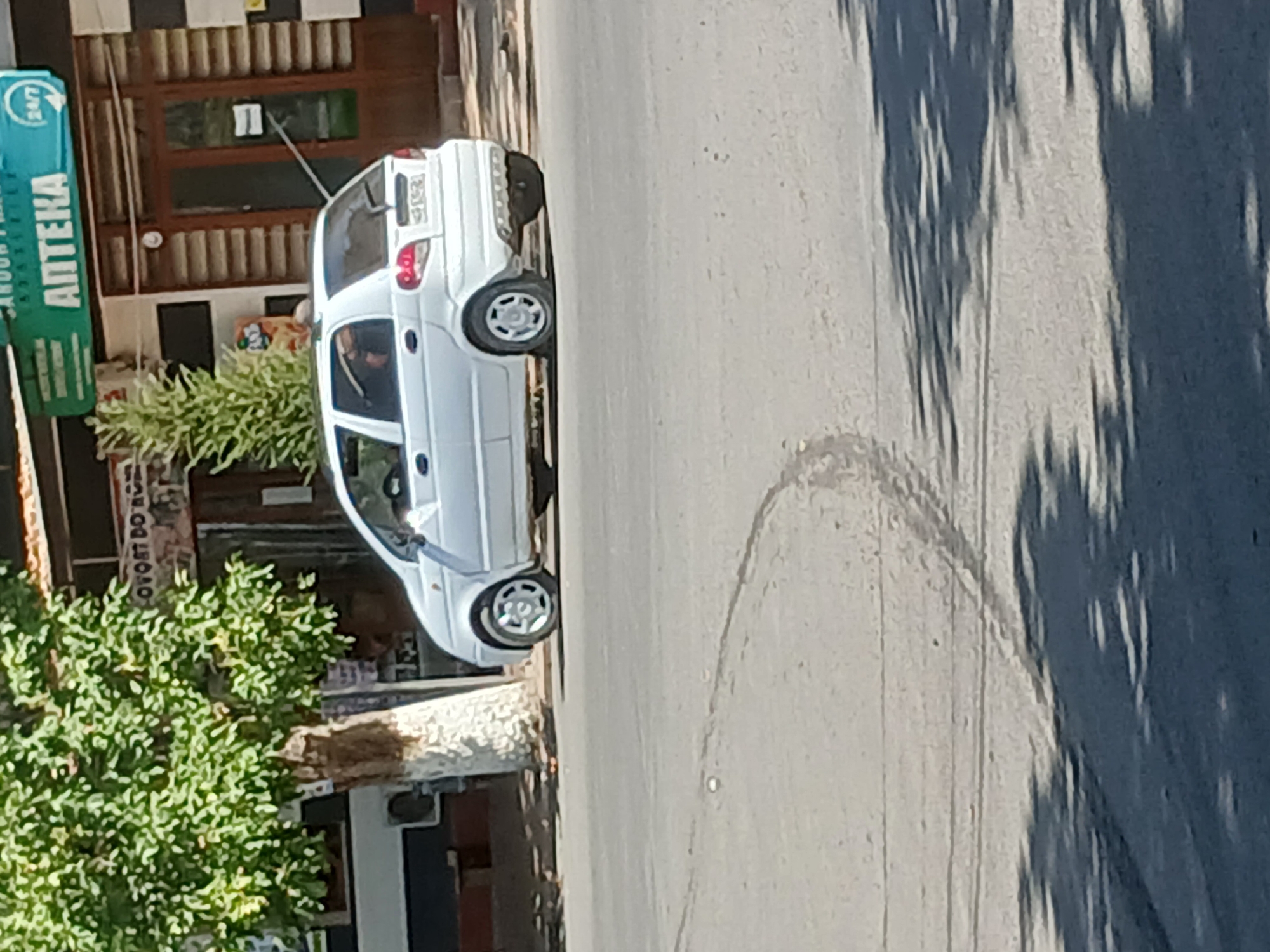 A white Daewoo Matiz is parked on the side of a street in Talmazar, Uzbekistan. The car is facing the right of the frame. The car's rear passenger-side tire is flat. The car is parked in front of a building with a green awning that reads "ANTEKA" and has a 24/7 symbol. There is a tree with green foliage to the left of the car. The street is paved, and there are tire tracks visible on the road. The lighting suggests it is daytime, with shadows cast on the road.