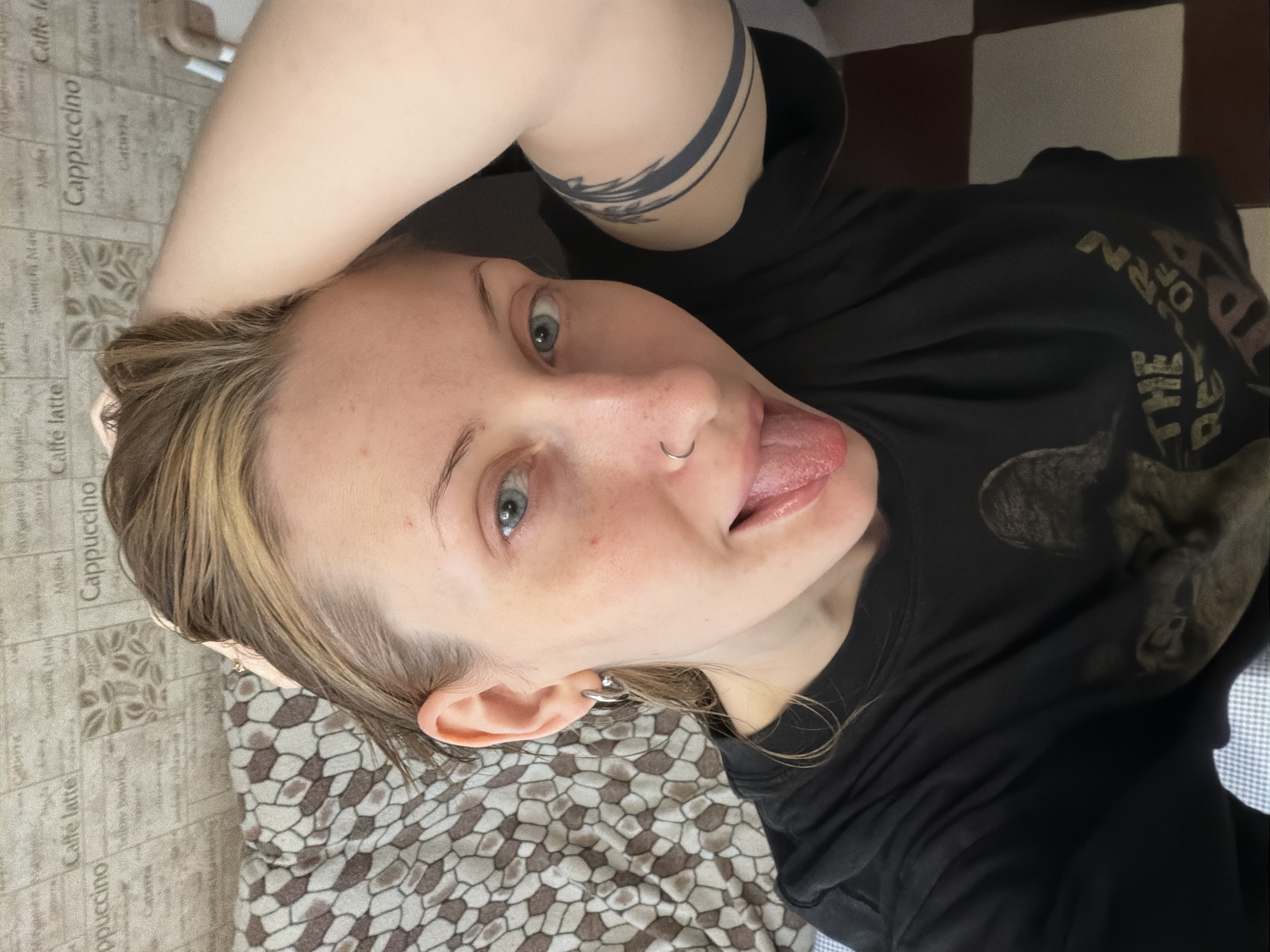 This is a close-up, overhead shot of a person lying down. They are wearing a black t-shirt with a graphic design on it, and have a tattoo on their left arm. The background includes a patterned blanket and a checkered floor. The person has their tongue sticking out and a nose ring. The overall mood is playful and casual. The scene appears to be indoors, possibly a bedroom, given the blanket and the relaxed pose. There is no indication of the time of day or weather conditions. The image does not contain any visible text. The location context provided is Saint Petersburg, Russia.