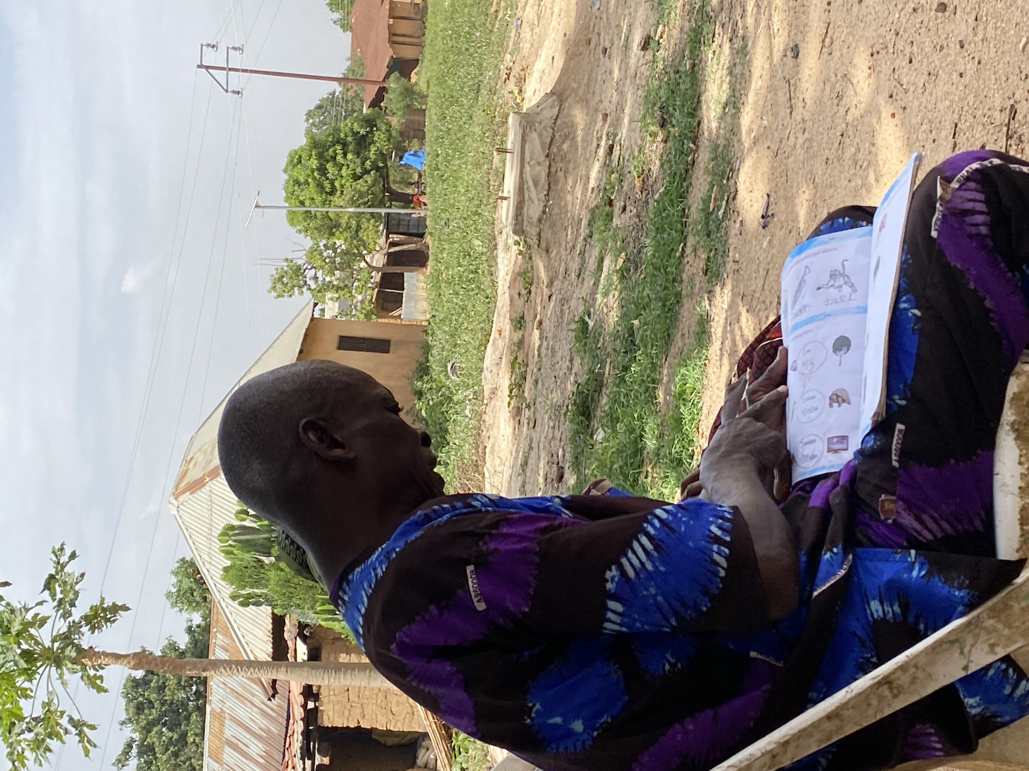 An elderly Black man sits in a white plastic chair outdoors in Kuru Dungus, Nigeria. He wears a blue and purple patterned shirt and dark pants, and a sheet of paper with hand-drawn illustrations and text rests on his lap, which he appears to be reading.
In the background, a rural landscape includes vegetation, dirt roads, and modest structures with corrugated metal roofing. Utility poles and wires are visible against the sky. A dirt road is partially visible through grass.