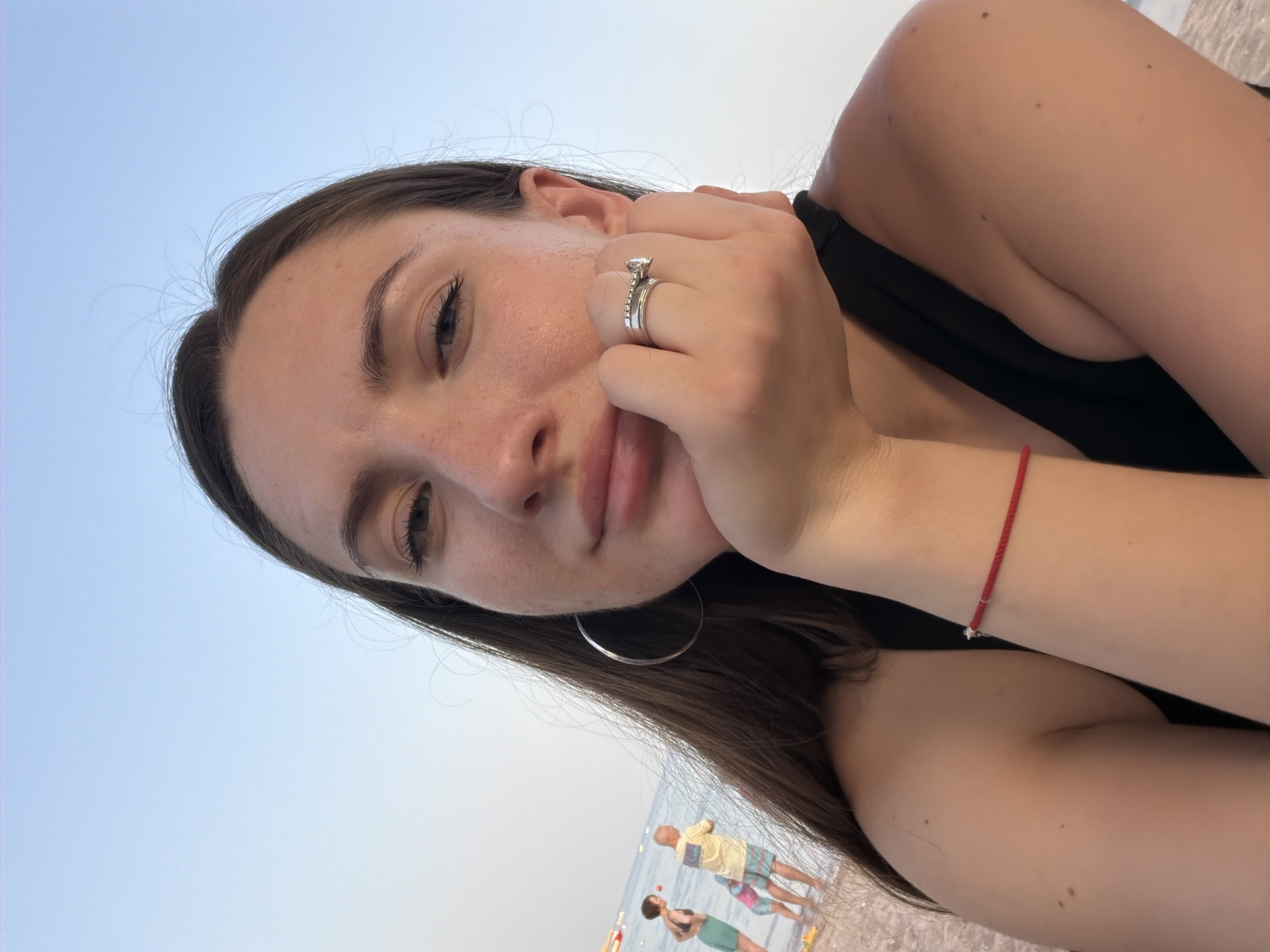 A close-up selfie features a young woman with long brown hair resting her chin on her hand. She is wearing a black top and a red string bracelet on her left wrist. A silver engagement ring is visible on her right ring finger. In the background, a beach scene unfolds with the sea and other beachgoers. The sky is clear and pale blue, suggesting it is late afternoon or early evening. The location is the beach in Sharm El Sheikh, Egypt.