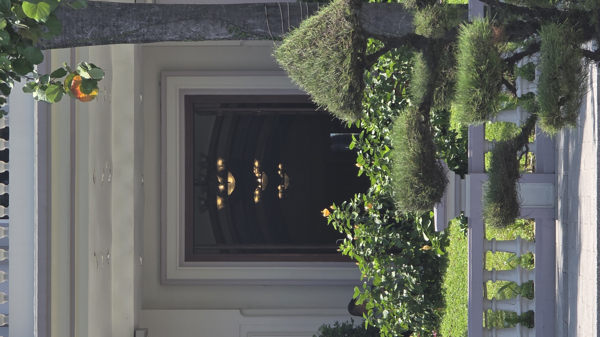 A slightly rotated, eye-level, exterior shot shows the entrance to what is likely a hotel or government building in Nha Trang, Vietnam. The building is a pale white, trimmed in pale gray. The doorway is large, set within a frame, and features chandeliers visible inside the dark interior. To the right, a precisely trimmed tree and fence are visible. The light is bright, appearing to be a sunny day. The scene is quiet and still. No people are clearly visible.