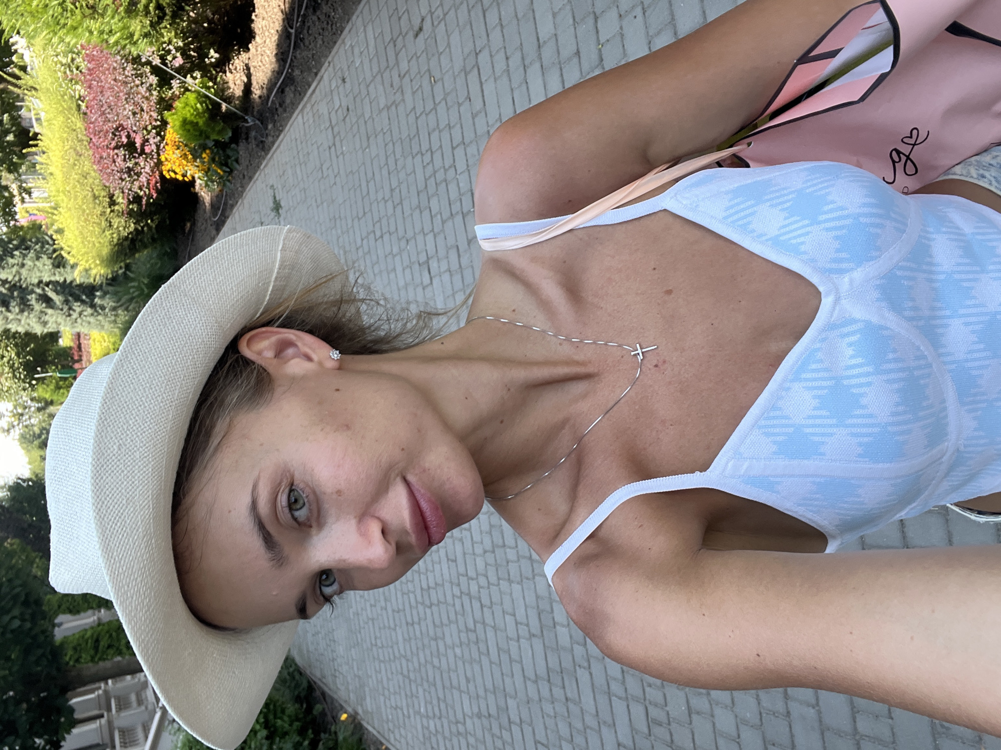 The image is a selfie taken by a fair-skinned woman with blonde hair, wearing a white wide-brimmed hat, a white top with a blue pattern, and a silver necklace with a cross pendant. She is also wearing a diamond stud earring. The woman is standing on a paved pathway outdoors, with a garden of colorful bushes and trees in the background. To her right, she is carrying a pink tote bag. The scene is consistent with a park or a garden area within the city of Lisky, Ukraine.