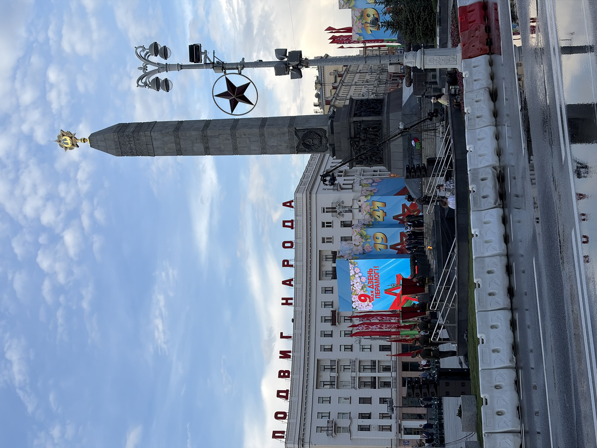 The image depicts a scene in Minsk, Belarus, likely during a commemoration of Victory Day (May 9th). The "Podvig Naroda" (Deed of the People) monument is prominently featured, topped with a gold star. A building is visible in the background with a banner displaying the text "9 May" and Cyrillic text, likely related to Victory Day. A line of soldiers is seen in formation in front of the building. A street with a temporary barrier is also visible in the foreground. The sky above is partly cloudy.