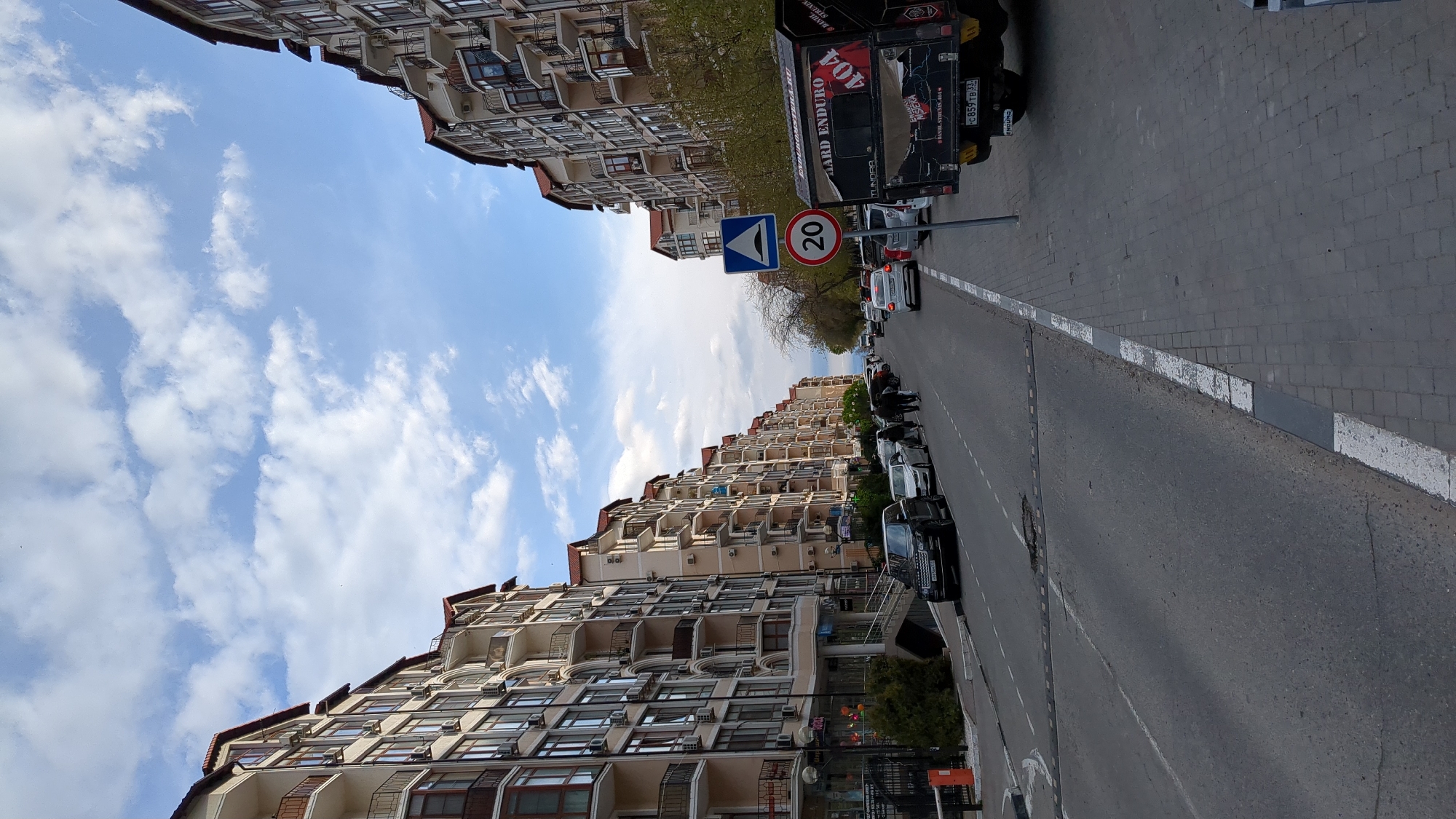 The image shows a street scene in Gelendzhik, Russia. The view is along a road lined with tall apartment buildings on both sides. The buildings have multiple floors, with many windows and balconies. Vehicles are parked along the right side of the road. A truck is parked further up on the right. A speed limit sign reading "20" is visible. The sky is blue with scattered clouds. The pavement is grey with white road markings along the edge. The truck has the license plate "C 859 TB 33". A sign on the truck reads "HARD ENDURO".