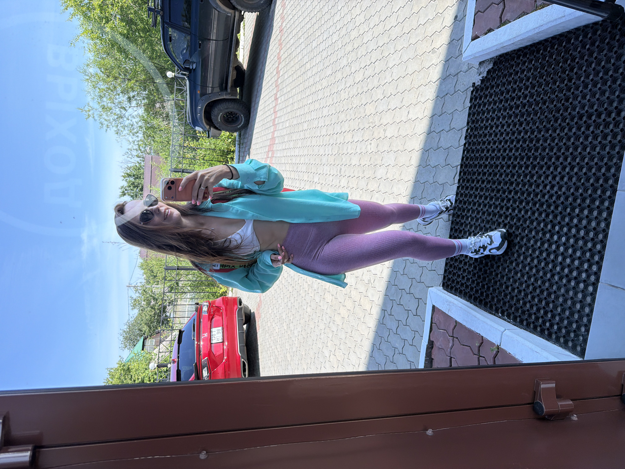 In Birobidzhan, Russia, a young woman is pictured in a full-body selfie taken in front of a building. She is wearing a white sports bra, a light blue open button-up shirt, and pink textured leggings. Her outfit is completed with white and black athletic shoes and white socks. She is also accessorized with sunglasses and a ring on her left hand, and she is holding a pink smartphone in her right hand, which she is using to take the selfie.
The background shows a paved area with a red car parked on the left and a dark colored SUV on the right. The sky is clear and blue, with some greenery visible in the distance. The overall scene appears to be outdoors, possibly in a residential or commercial area.