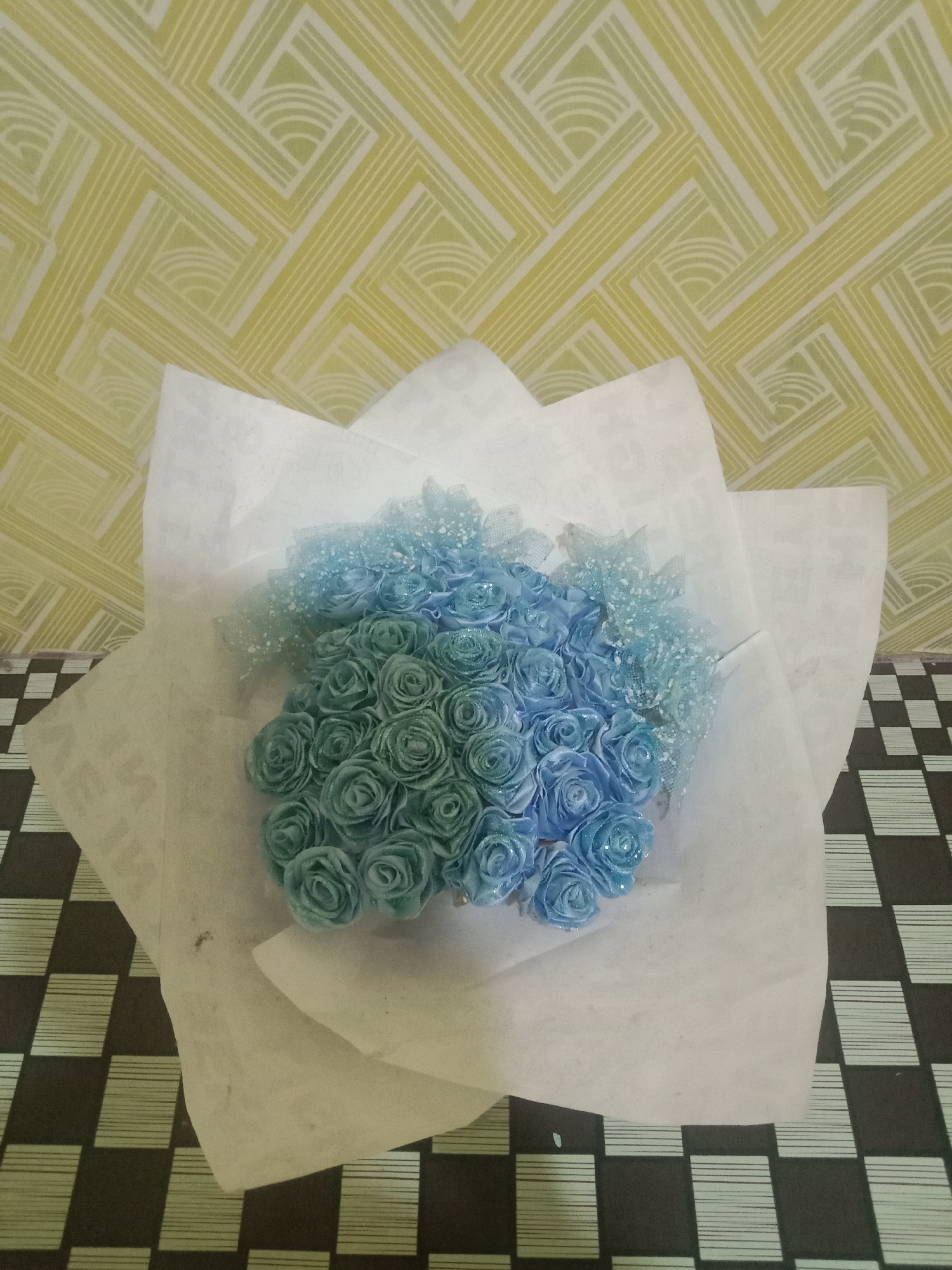 A bouquet of blue and teal roses, some with glitter, is presented on a surface with a black and white checkered pattern. The background features a wall with a geometric, green and yellow pattern. The image is a still life, with no people or actions depicted. The location is not specified in the image or its context.