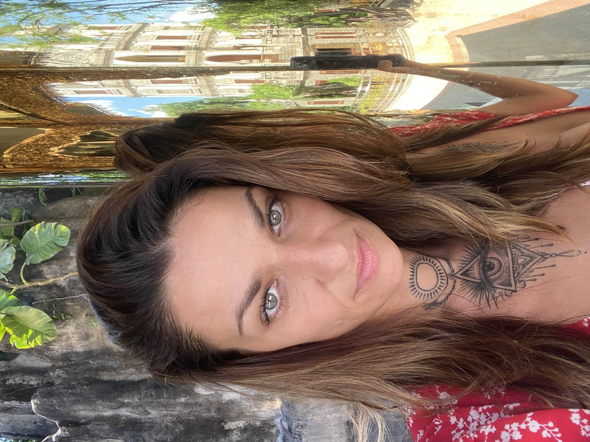 The image is a selfie of a woman, likely taken in Dĩ An, Vietnam, given the architectural reflection above her. She has light eyes and shoulder-length brown hair. A tattoo is visible on her chest and neck. She is wearing a red floral dress. The background includes weathered stonework and foliage, as well as a reflection of a building with intricate details and possibly a garden. The lighting is bright and natural. She is holding a phone to take the picture.