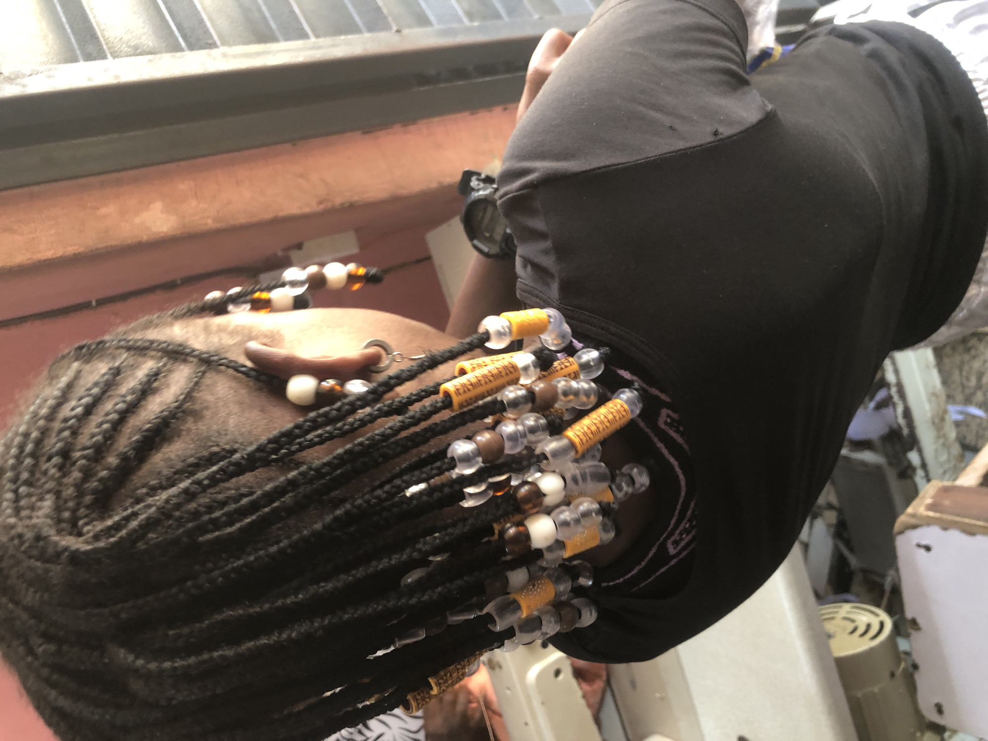 The image depicts a close-up of a young person's braided hair, adorned with beads. The braids are dark and appear to be styled in cornrows, with individual braids adorned with various colored beads including white, brown, clear, and yellow. Some of the yellow beads have a cylindrical shape with etched patterns. The person is wearing a black t-shirt. In the background, a portion of a wall painted in shades of orange and brown is visible, along with a metal grate or blinds. A sewing machine is partially visible on the left side of the image, suggesting an indoor or workshop environment. The lighting suggests it is daytime. The image does not contain any explicit information about the location being Ado, Nigeria, nor any specific details of the surrounding environment that would definitively place it in that city.