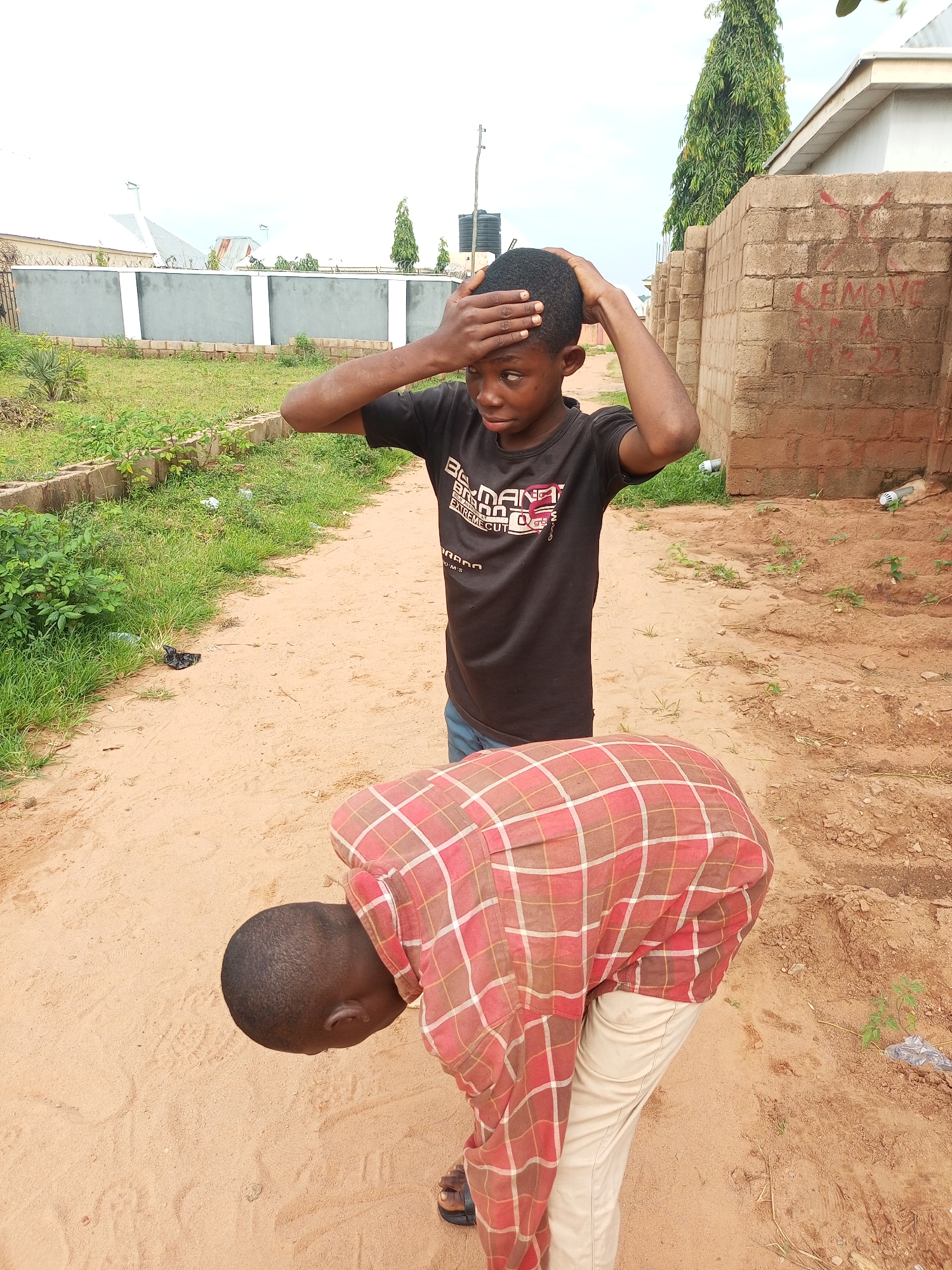In Tempure, Nigeria, two young boys stand on a dirt path. The boy in the foreground, wearing a red and brown plaid shirt and tan pants, is bent over with his back to the camera. The boy in the background, facing the camera, wears a black t-shirt with a logo and blue jeans. He has his hands on his head and appears to be experiencing distress. The environment is an outdoor setting with a dusty path, green grass, and a brick wall with writing. A satellite dish and buildings are visible in the distance.
