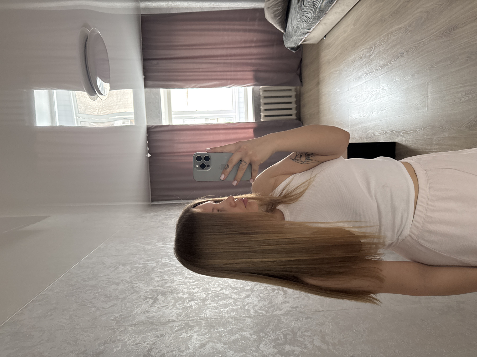 A person is taking a selfie in a room. The room appears to be a bedroom with a bed and window visible. The blinds are drawn. A radiator is next to the window. The walls and floor are light colored. It is not possible to confirm the location as Chaikovsky, Russia with the current information.