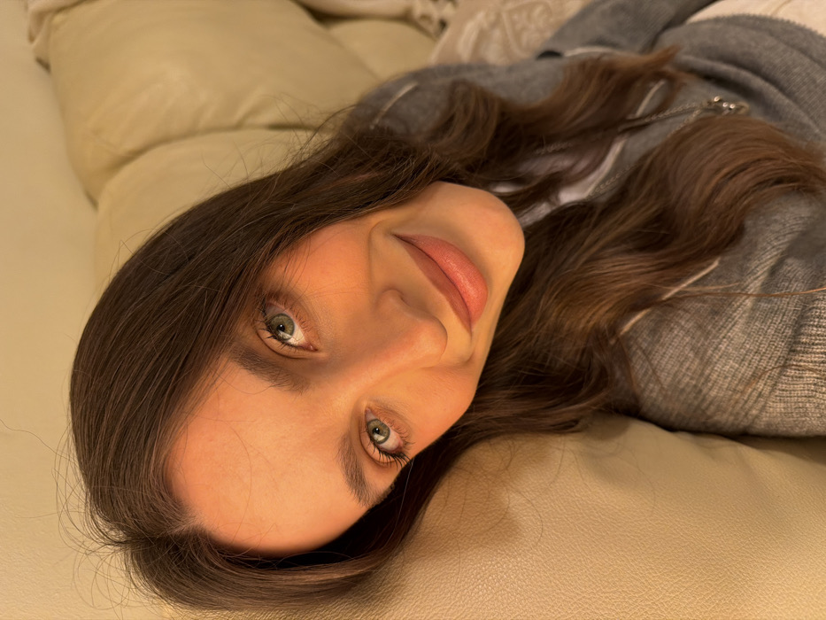 A young woman with long, wavy brown hair and fair skin reclines on a light beige couch. Her green eyes are visible, along with a gentle smile and full lips. She's wearing a gray sweater with white trim. The background consists of the couch and a white textured blanket or pillow. The image is slightly rotated, presenting a close-up view of her face.