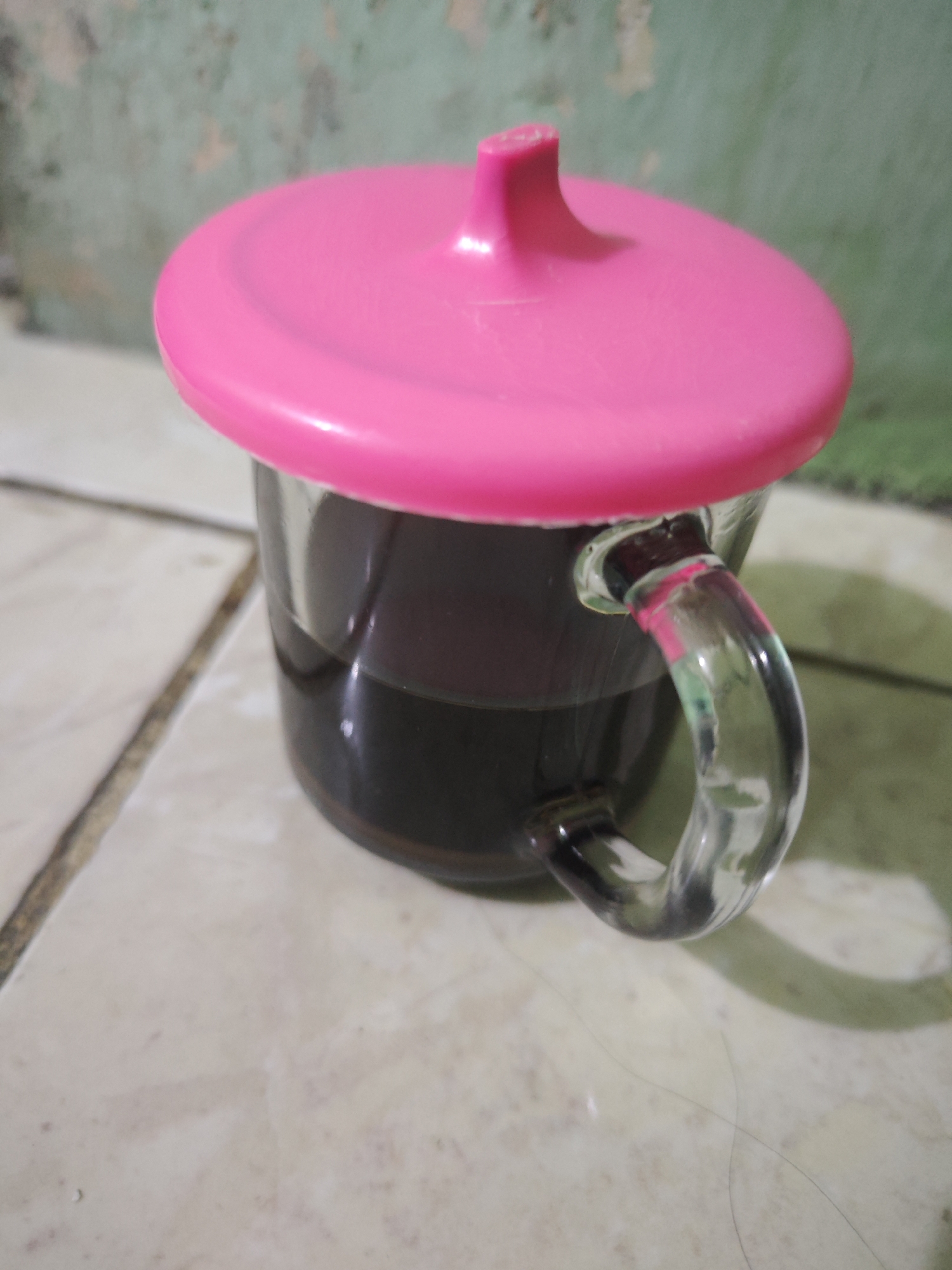 The image shows a close-up, eye-level shot of a clear glass mug filled with a dark liquid, likely coffee, and topped with a bright pink plastic lid. The mug has a transparent, curved handle. The mug is placed on a tiled surface, possibly a floor, with visible grout lines. In the background, a wall with a distressed, mottled green and white paint finish is visible. The scene appears to be indoors, possibly in a home or a small establishment, in the city of Sroyo, Indonesia. No people are present in the image.