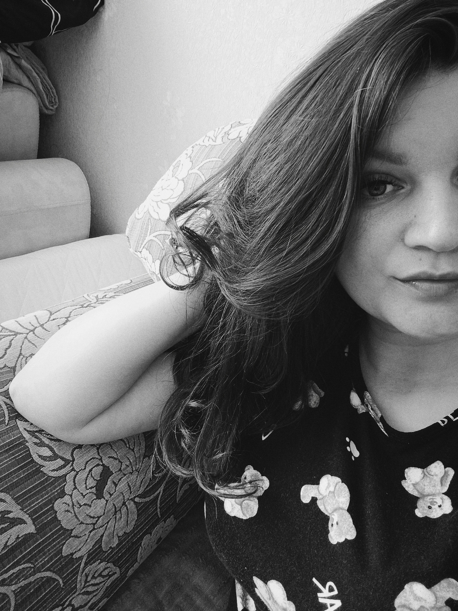 The image is a black and white close-up of a woman. She has long hair that falls over her face and shoulder. She is lying down, her head resting on a pillow with a floral pattern. Her arm is bent and resting on the pillow as well. She is wearing a dark top with a pattern of teddy bears. Only part of her face is visible, but she appears to be looking at the camera. A portion of a couch is visible in the background, along with a suggestion of a wall behind it. There is no evidence to suggest that this image was taken in Tobolsk, Russia.