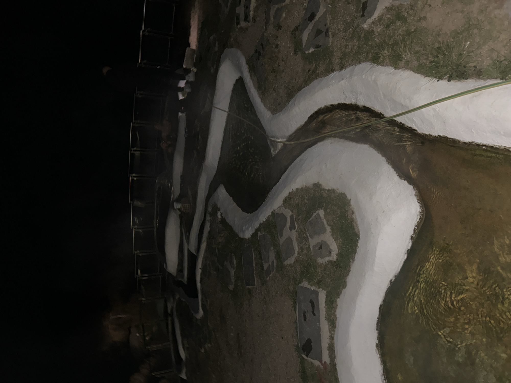 The image shows a winding artificial water feature, likely a stream or canal, with white-painted borders. The water within appears murky and brown. A green hose lies across the water feature. The surrounding area consists of patchy grass, dirt, and several rectangular paving stones. To the left of the frame is a dark, vertical structure that appears to be a metal staircase or railing. The overall lighting is dim. The location is in Iamatikes Piges, Greece.