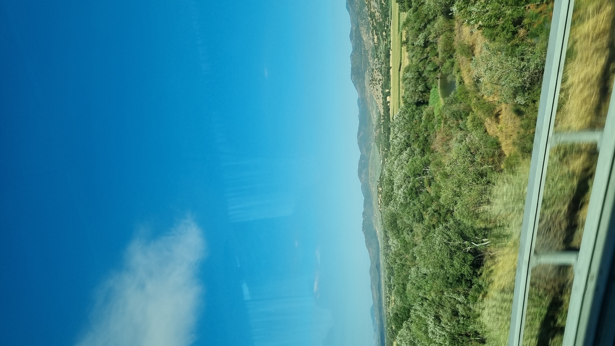 The image captures a serene rural landscape under a clear blue sky with a single white cloud on the left. The foreground features a highway guardrail with a blurred effect indicating movement, suggesting the photo was taken from a vehicle. Beyond the guardrail, a dense expanse of lush green trees and vegetation stretches into the middle ground. A small, dark body of water is visible within this green expanse, possibly a pond or a small lake. In the background, rolling hills and mountains recede into the distance, their forms softened by atmospheric perspective. The scene is likely located in the Municipal Unit of Makrichori, Greece, a region known for its picturesque natural beauty and undulating terrain. There are no people or animals visible in the image.
