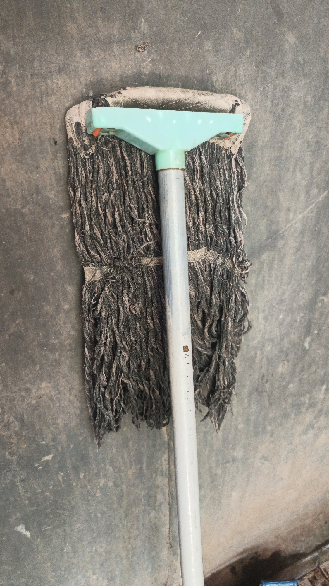 A mop with a light blue plastic head and a silver handle lies on a rough, gray concrete surface. The mop head is made of dark gray fabric strands, which are tied together with a lighter colored band. The strands appear slightly frayed and dusty. The concrete surface has some dirt and scuff marks. A portion of a dark area, possibly a shadow or a drain, is visible at the bottom right of the image. The scene appears to be an indoor or covered outdoor space, as indicated by the lighting and the concrete flooring. The image is taken from a top-down perspective.