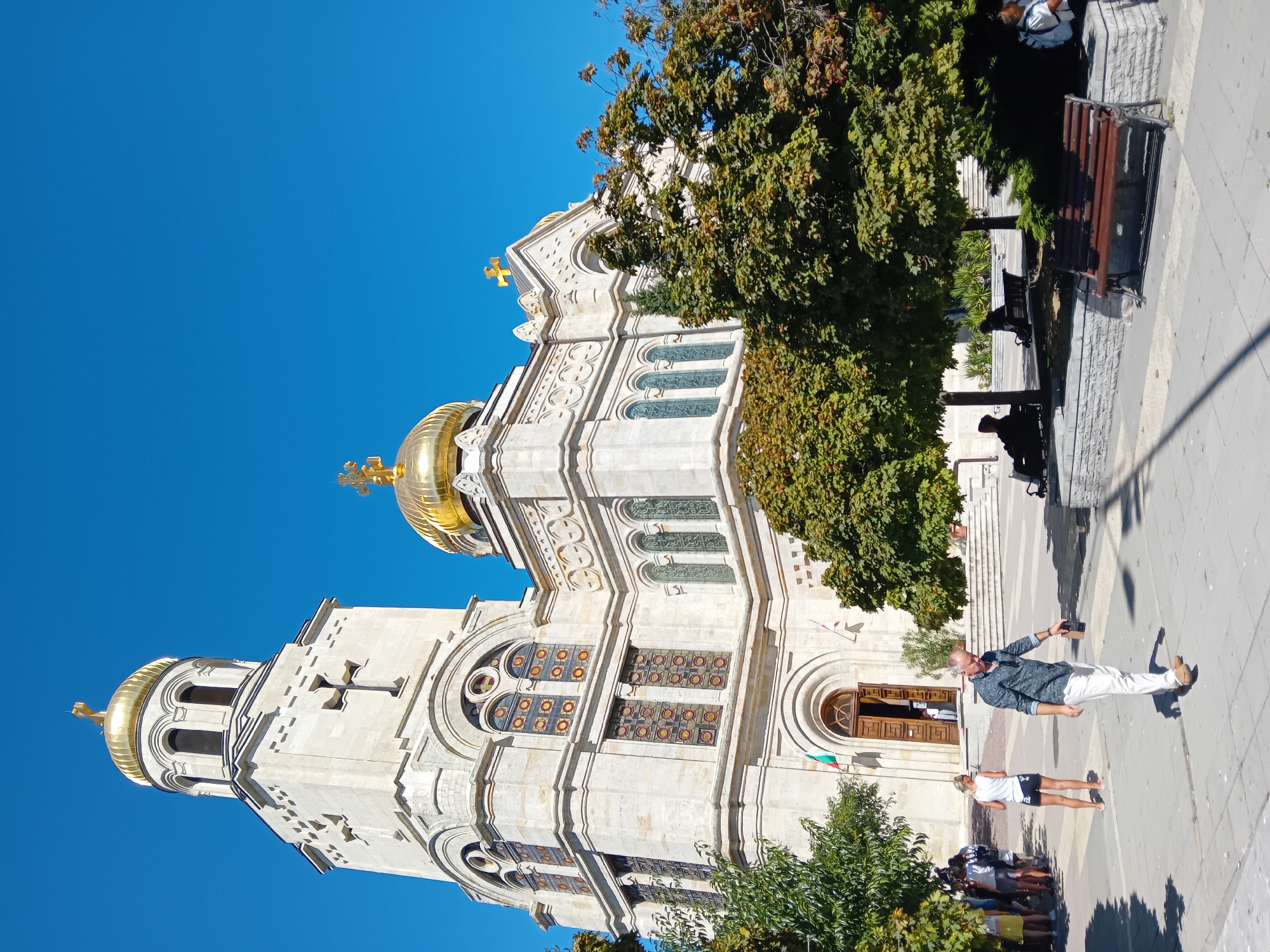 A wide-angle, eye-level shot captures the imposing facade of the Cathedral of the Assumption of the Virgin Mary in Varna, Bulgaria, on a bright, sunny day with a clear blue sky. The cathedral, built of light-colored stone, features a prominent bell tower on the left, topped with a golden dome and cross, and a central main structure with a larger golden dome and cross. Intricate architectural details, including arched windows with stained glass and decorative carvings, are visible.
In the foreground, a paved plaza leads to the cathedral entrance. Several people are present: a man in a blue patterned shirt and white pants walks towards the right, looking down and holding a dark object in his right hand. To his left, a woman in a white tank top and black shorts stands facing away from the camera. Further left, a group of people are gathered, some with backpacks. Several park benches are scattered around the plaza, with some individuals seated on them, casting shadows from the surrounding trees. Lush green trees with full canopies are strategically placed in front of and around the cathedral, providing shade and adding to the park-like atmosphere of the plaza. A Bulgarian flag is visible near the cathedral entrance.