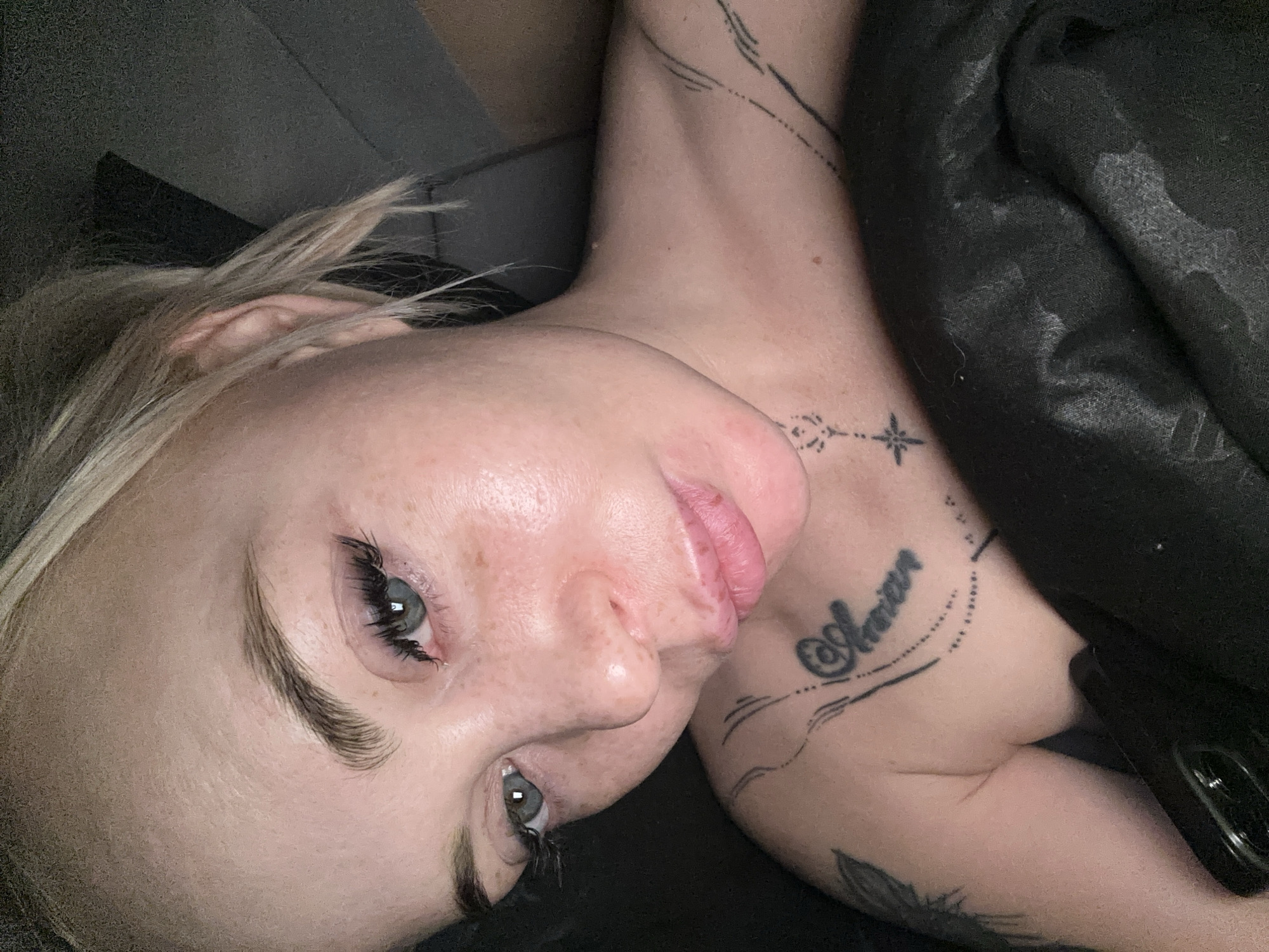 The image presents a close-up, high-angle portrait of a person, likely female, with blonde hair, light skin, and blue-grey eyes, lying down. The person's head is tilted, creating a rotated perspective within the frame. Freckles are visible across the nose and cheeks, while the lips are full, pink, and show some natural texture. Long, dark eyelashes enhance the eyes, and the eyebrows are well-defined.
Several tattoos are prominent on the neck and upper chest. These include delicate dotted lines and a star-like symbol near the collarbone. A script tattoo on the chest appears to spell "Aarian" or a similar name, accompanied by other linear designs.
The person is partially covered by a dark, textured fabric, possibly a blanket or duvet, which shows some lint or wear. A dark-colored smartphone, with its camera module visible, rests beside the person's arm on the right side of the frame.
The background is dark and indistinct, suggesting an indoor setting such as a bedroom or living area. The lighting is soft and somewhat dim, contributing to a relaxed atmosphere. The person's expression appears neutral or contemplative, with a slightly parted mouth. No specific time of day is discernible, but the dim lighting suggests a low-light environment or evening hours.