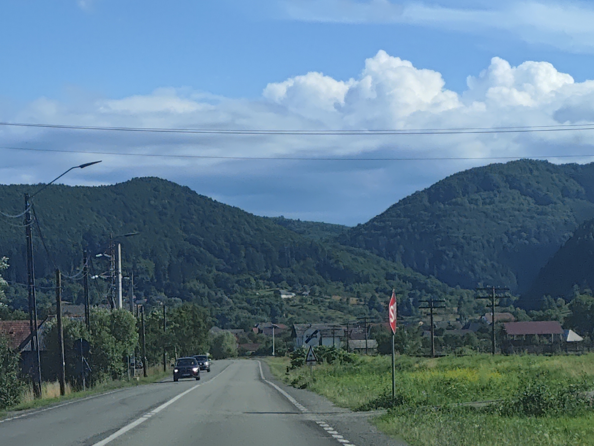 The image depicts a rural road scene in Leordina, Romania. A two-lane asphalt road curves slightly to the right as it approaches a cluster of buildings nestled at the base of densely forested, rolling hills. Two cars, a dark SUV and another dark vehicle, are traveling in the same direction as the camera. Along the left side of the road, there are utility poles with wires, streetlights, and some residential structures partially obscured by trees. On the right side of the road, a red and white stop sign is prominently displayed on a pole, accompanied by a triangular warning sign below it. Beyond the road and signs, fields of green grass and some yellow wildflowers are visible, leading up to the village buildings and the imposing green mountains in the background. The sky is a vibrant blue, punctuated by large, fluffy white clouds and some darker, more diffused cloud formations, suggesting a partly cloudy day. The overall impression is one of a serene, pastoral setting within the Romanian countryside.