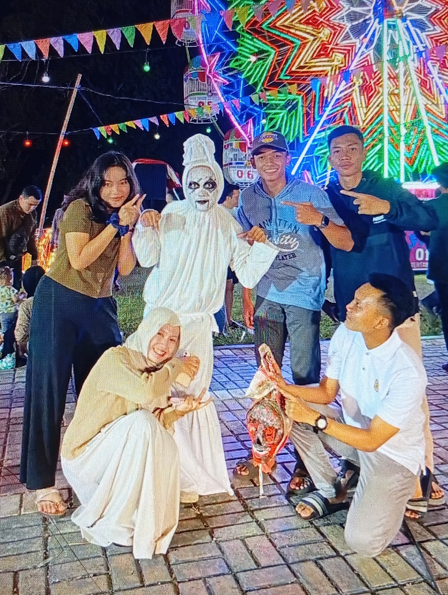 The image depicts a group of people posing for a photo at night, with a brightly lit Ferris wheel in the background, suggesting a fairground or amusement park setting. Several individuals are present, including a person in a white ghost costume, two young women, and three young men. One woman, wearing a dark top and black pants, is on the left, gesturing towards the ghost. Another woman, dressed in a light-colored hijab and dress, is crouching in the foreground, smiling and holding a small, possibly decorative, object that resembles a stylized skull with colorful adornments.
Two young men stand behind the crouching woman. One is wearing a blue hooded sweatshirt and a cap, pointing at the ghost. The other, in a black long-sleeved shirt, is also pointing. A third young man, in a white polo shirt, is kneeling to the right, looking towards the object held by the crouching woman and gesturing with his hand.
The environment is paved with interlocking bricks, and festive banners are strung across the scene. The overall atmosphere suggests a lively event, possibly a celebration or a local festival. The presence of the ghost costume and the decorative skull-like object hints at a Halloween theme or a similar cultural event. Based on the information available, the scene is likely located at a fairground in Lhokseumawe, Indonesia, possibly during a local festival or event.
