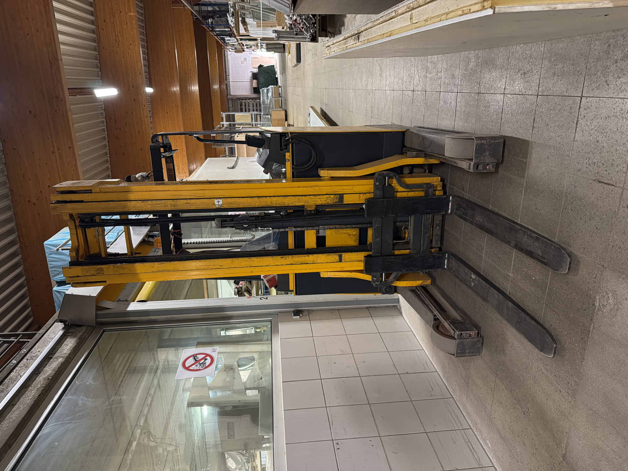 The image shows a yellow and black forklift inside a building in Leudelange, Luxembourg. The building has tiled floors and walls. The forklift has two metal forks extending forward. A sign with a crossed-out cell phone symbol is visible on a window near the forklift. The building appears to be an industrial or commercial space with wooden beams and metallic elements in the ceiling.