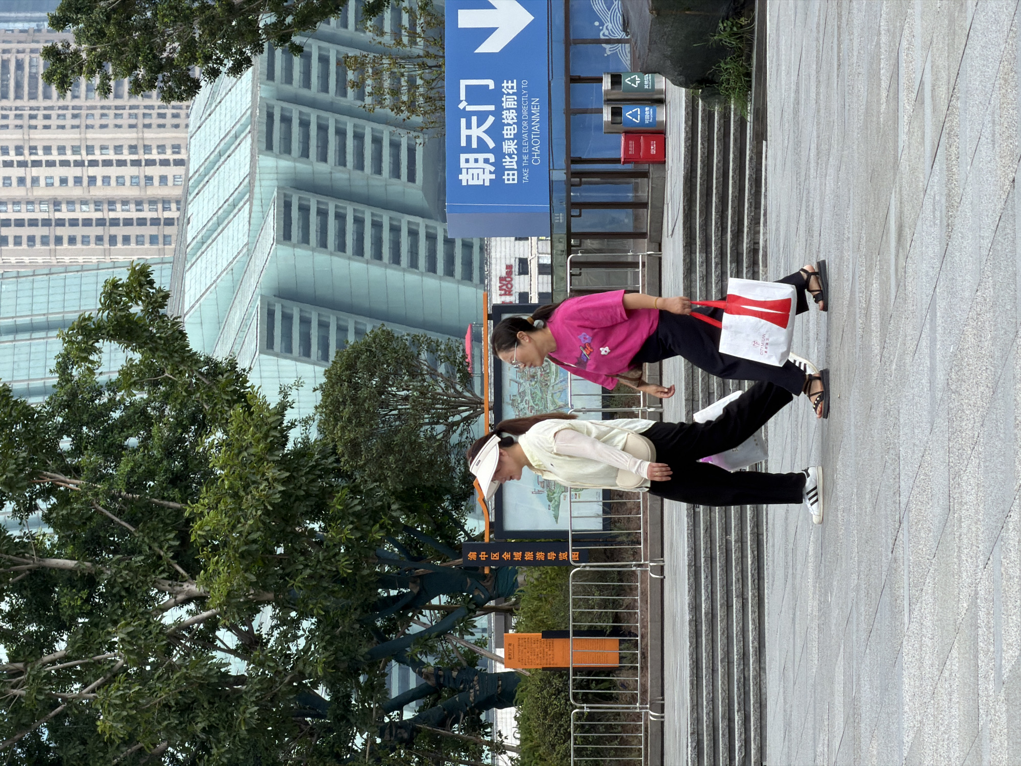 In Yuzhong District, China, two women walk on a paved plaza in front of a modern building with a unique blue glass facade. The woman on the left, wearing a light-colored long-sleeved shirt, black pants, and white sneakers, carries a white shopping bag. She has a white visor on her head and a small purse slung over her shoulder. The woman on the right is dressed in a bright pink t-shirt with a floral design, dark pants, and dark sandals. She is carrying a white shopping bag with red accents. Behind them, a blue sign with white text provides directions in Chinese and English, indicating "TAKE THE ELEVATOR DIRECTLY TO CHAOTIANMEN." Several trees with green foliage are scattered throughout the scene, and further in the background, tall office buildings are visible. Two trash receptacles, one red and one green, are positioned near the blue sign. The ground is wet in some areas, suggesting recent rain.
