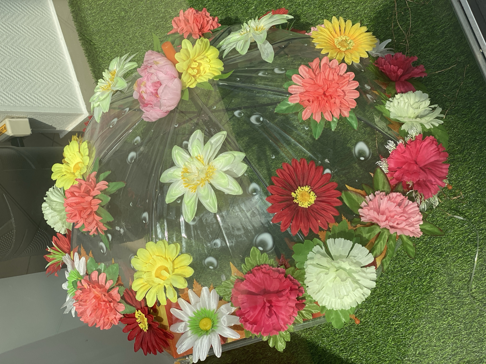 This is a photograph of an umbrella decorated with artificial flowers. The umbrella is made of clear plastic and is open, lying on a surface of artificial green grass. Various artificial flowers in different colors, including pink, red, yellow, white, and green, are attached to the umbrella's canopy and spokes. Some flowers have many thin petals, while others are more rounded and ruffled. The umbrella appears to be displayed outdoors, as indicated by the artificial grass and the background, which includes a textured wall and a light-colored structure. There are no people visible in the image, and no specific location in Novokuznetsk, Russia can be identified from the provided visual information.