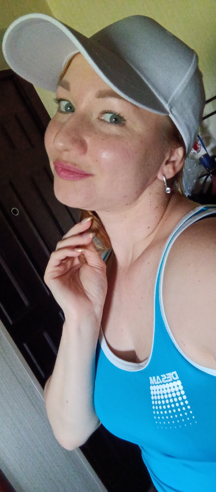 A woman with green eyes, freckles, and pink lipstick is wearing a light grey baseball cap and a blue tank top with a white trim and a design that includes white circles and lettering, posing indoors. She has gold nail polish on and is wearing a dangling earring with a small stone. Her hand is near her face in a thoughtful pose. A dark brown door is visible in the background to the left. The exact location is unknown.