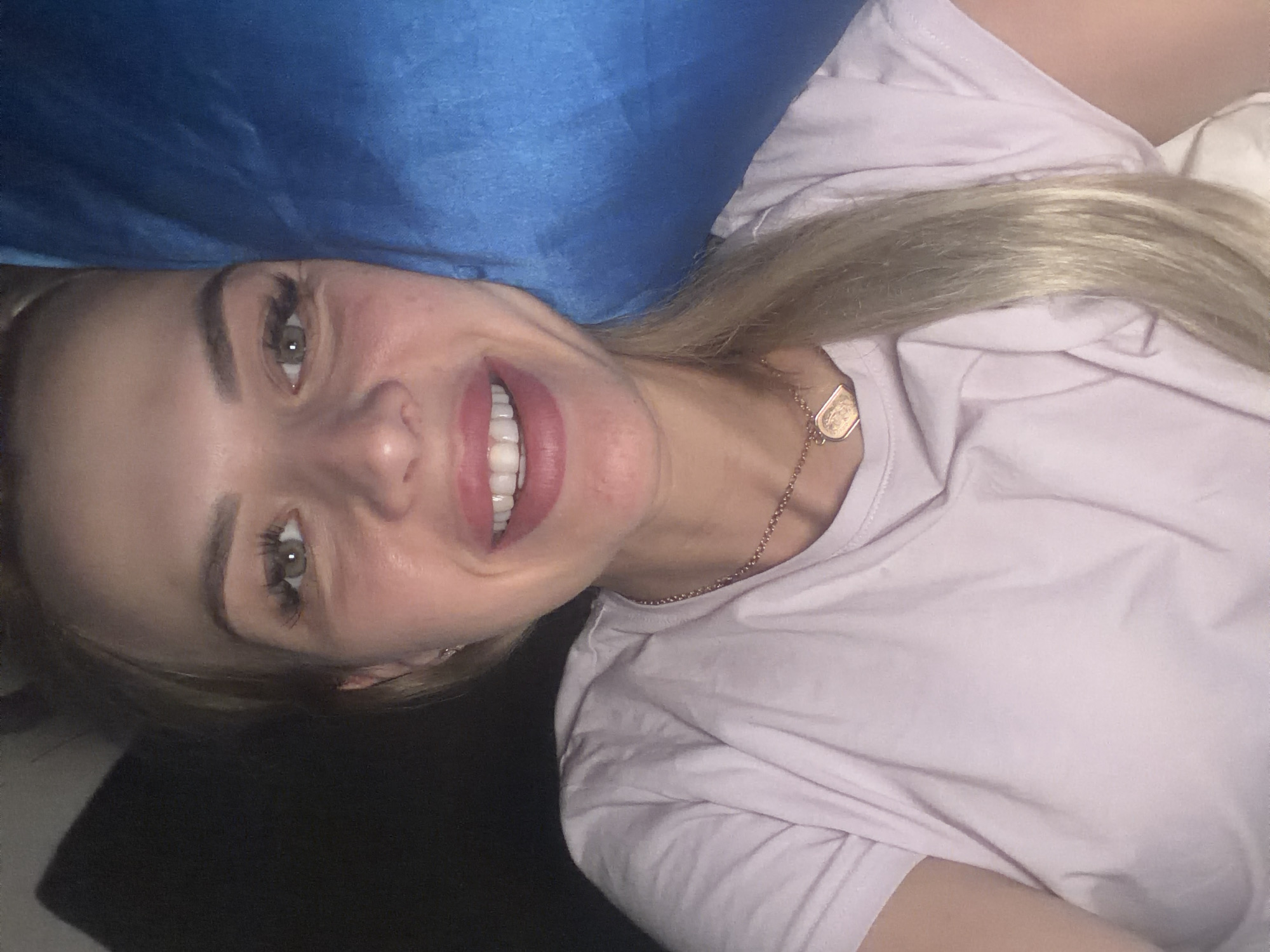 The image is a close-up selfie of a young woman with blonde hair and green eyes, smiling and looking directly at the camera. She is wearing a light purple t-shirt and a gold necklace with a pendant. She has her hair pulled back into a ponytail, which is visible on the right side of the image. The background is dark, with a hint of a blue object on the right, possibly a pillow or blanket. The image appears to have been taken indoors, in a dimly lit environment. There are no specific indicators of the location being in Kyiv, Ukraine.