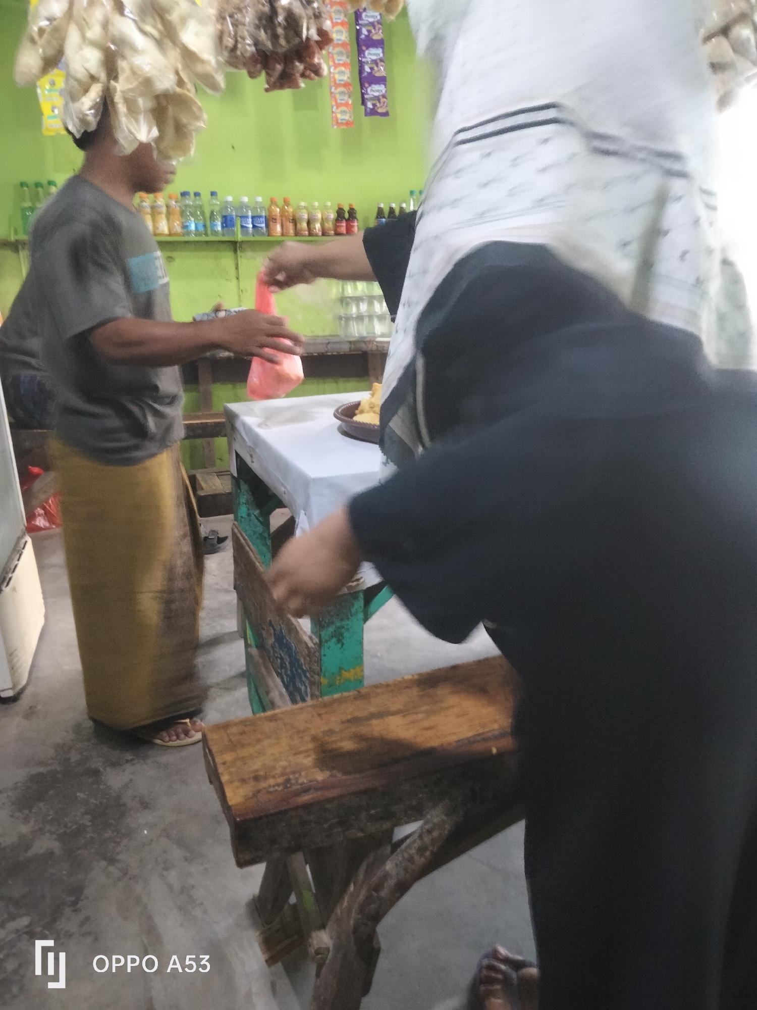 In Badung, Indonesia, a market scene unfolds. A person wearing a grey t-shirt and a long yellow sarong stands to the left, reaching out to hand a red plastic bag to another person whose torso is visible on the right, clad in black clothing and a white headscarf. They are standing near a table covered with a white cloth, with a brown bowl containing food on it. Behind them, shelves are stocked with various bottled drinks and packaged goods. The environment appears to be an open-air stall or small shop.