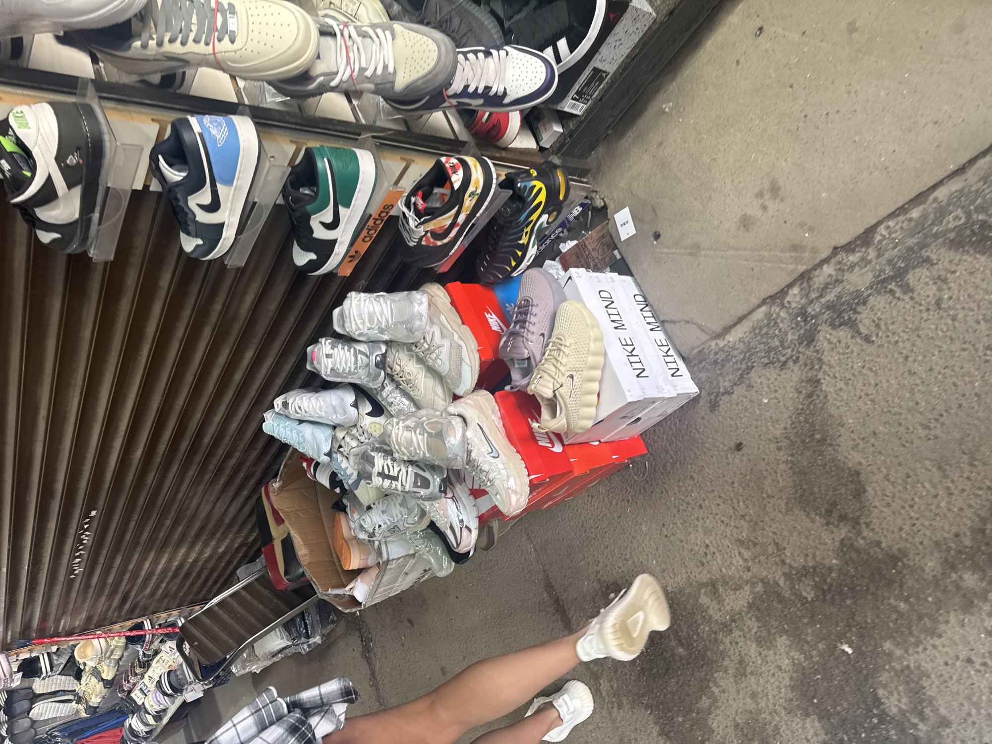 The image depicts an outdoor market stall in Saint Petersburg, Russia, specializing in the sale of athletic footwear. The stall features numerous pairs of sneakers displayed on shelves and stacked in boxes. A person, partially visible on the left, is wearing white athletic shoes and standing on a damp concrete surface. The background shows a corrugated metal shutter, a common feature in urban markets. The overall scene suggests a bustling commercial environment, possibly a street market or a temporary retail setup.
