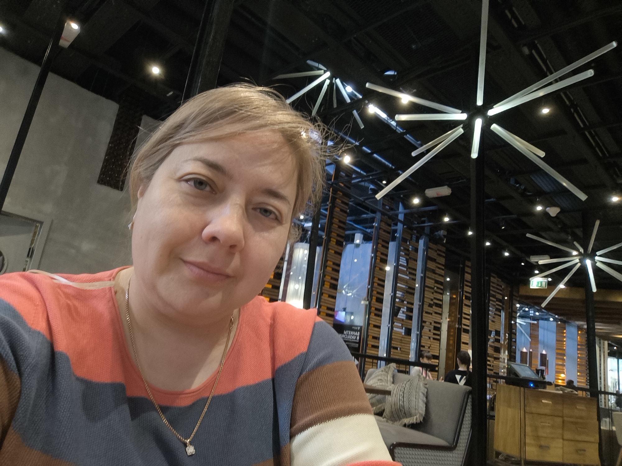 A woman is taking a selfie in what appears to be a restaurant or café in Khimki, Russia. She is wearing a striped sweater and a gold necklace. The background features modern lighting fixtures shaped like stars, a wooden architectural design element on the walls, and other patrons in the seating area. The furniture is contemporary, including upholstered chairs and wooden cabinets.