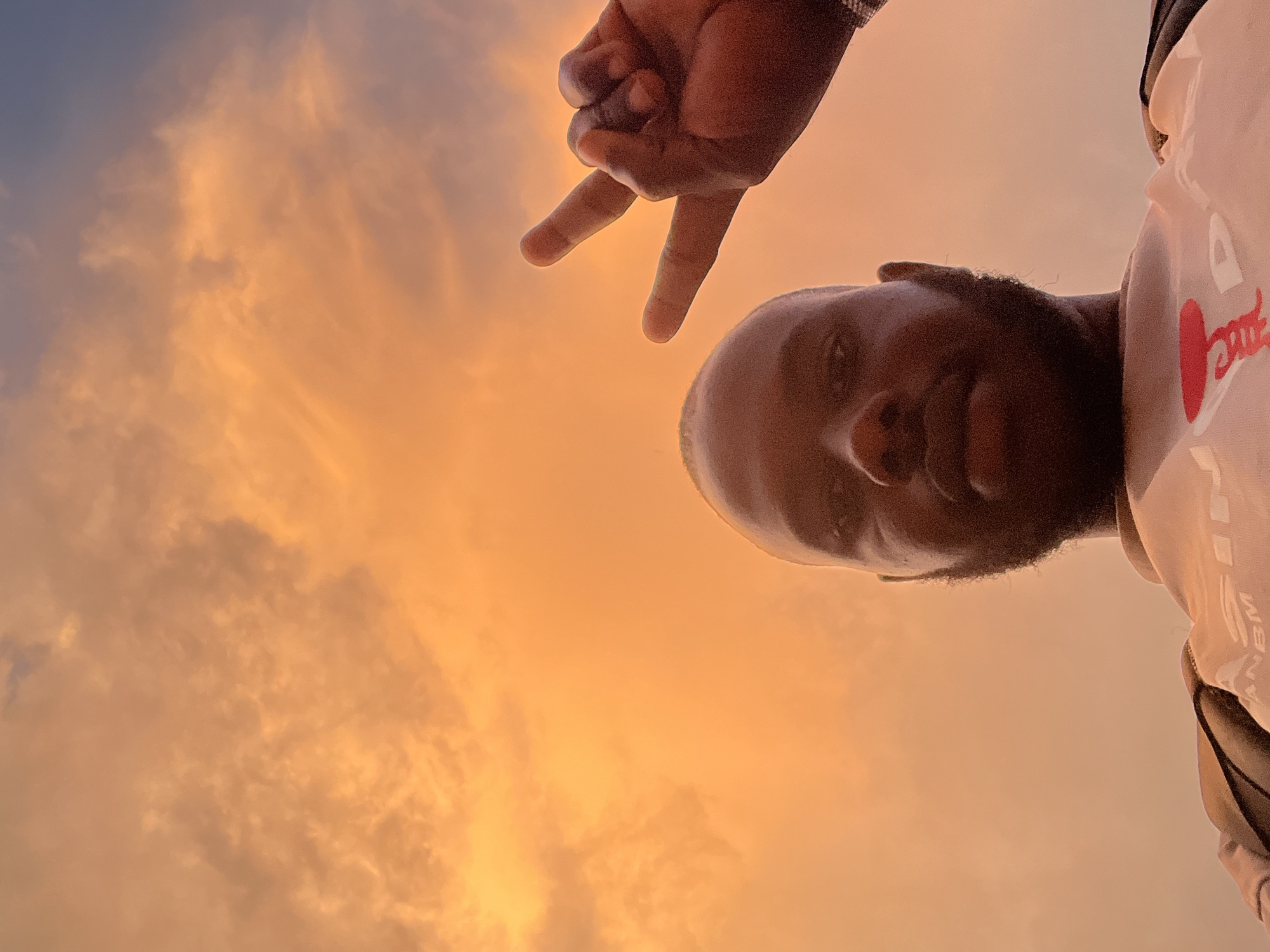 This image, rotated 90 degrees counter-clockwise, features a dark-skinned person's face and hand against a vibrant sky, set in Jos, Nigeria. The person's head, with a short beard, occupies the right side of the frame, looking directly towards the viewer. Their right hand, extended from the upper left, forms a "peace sign" gesture, positioned above their head relative to the natural orientation of the subject.
The individual wears a light-colored top, on which visible text reading "ANBM" can be seen on the sleeve, and a red heart-shaped graphic with cursive text is partially visible on the chest. The expansive background is a dramatic sunset or sunrise sky, characterized by warm hues of orange, yellow, and soft pink, indicative of the golden hour. Numerous scattered clouds, ranging from wispy formations to denser patches, are illuminated by this golden light, with some areas exhibiting cooler blue-grey tones. The scene captures a serene outdoor moment, using the rich, glowing colors of the sky as a striking backdrop for the portrait.