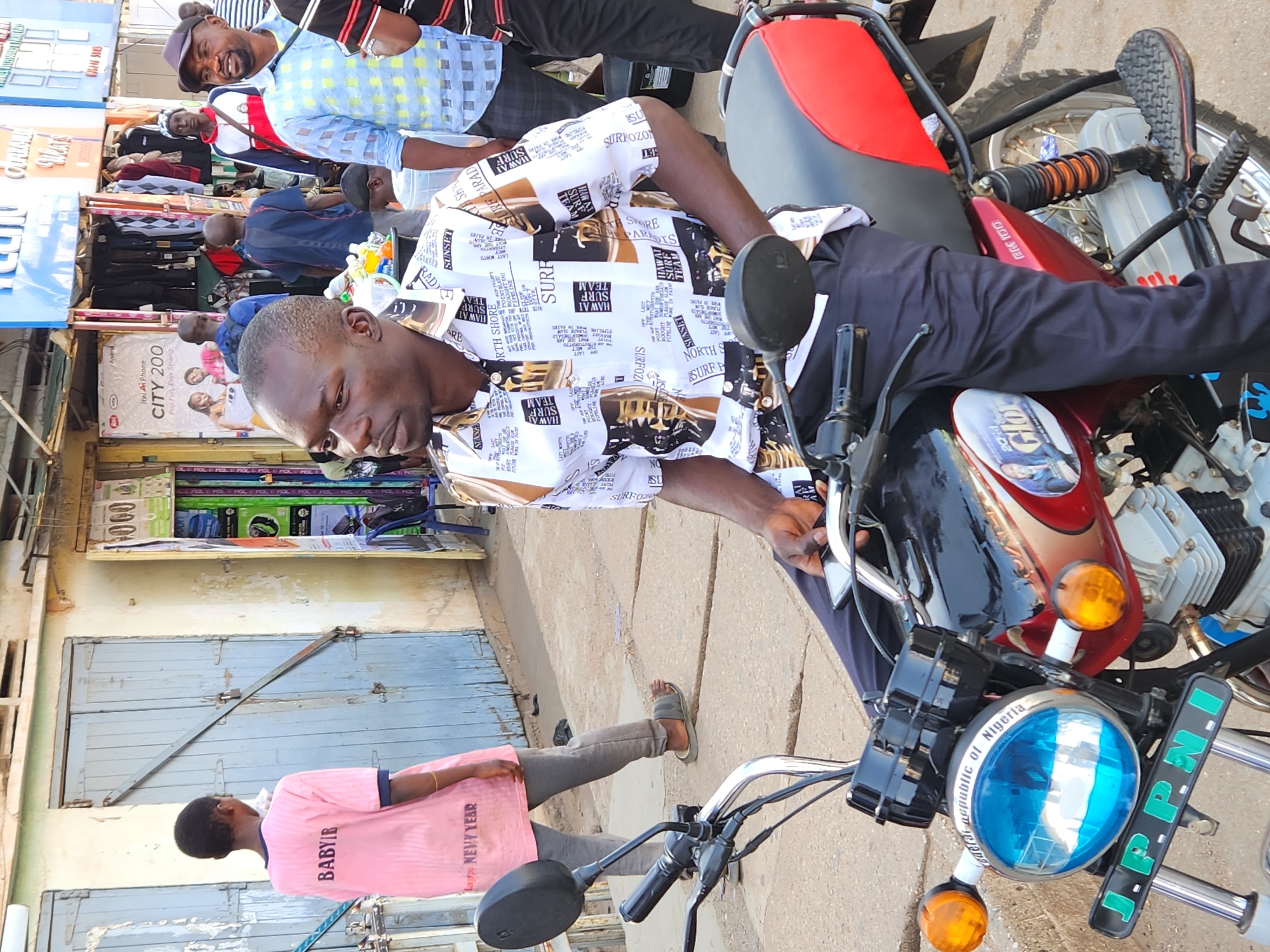 The image shows a man sitting on a red and black motorcycle in what appears to be a commercial street in Jos, Nigeria. The motorcycle has a license plate that reads "Federal Republic of Nigeria." The man is wearing a white shirt with black and brown print, and black pants. Behind him are several other people, including one man wearing a blue checkered shirt. A sign for "itel Ai Phone CITY 200" is visible. The street appears to have shops selling clothing. A person wearing a pink shirt that says "BABYIB" is walking away from the camera.