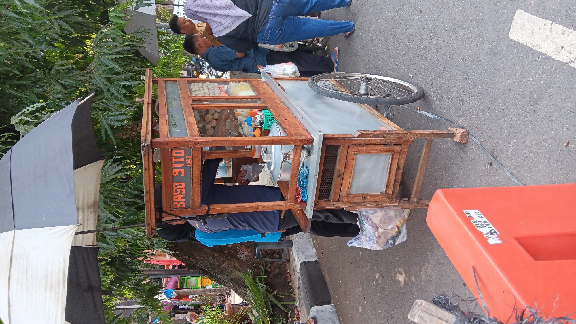 A street scene in South Jakarta, Indonesia, captures a meatball cart under a large black and white umbrella. The cart, made of wood and metal, displays the text "BASO SUTO 1922" on its front. A large metal pot, presumably containing meatballs, sits on the counter. Several people are gathered around the cart. A person in a blue shirt and headscarf stands to the left of the cart, facing away from the camera, holding a plastic bag. Two men and a woman in a light purple headscarf are on the right side of the cart, with one man interacting with the cart. A large orange traffic barrier with a sticker reading "DEFEND THE POLICE!" is in the foreground on the left. The scene takes place on an asphalt road, with trees and foliage visible in the background.