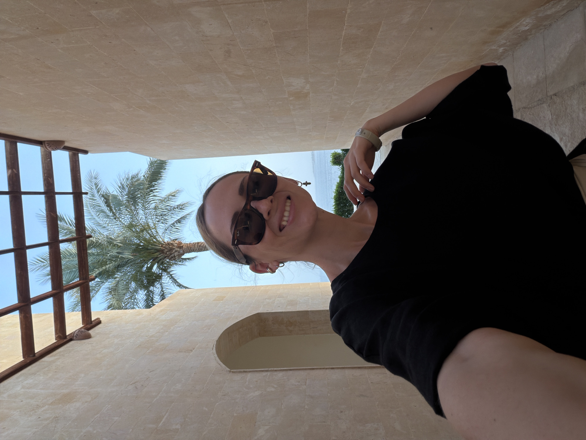 A woman wearing sunglasses and a black shirt stands in a beige-colored building with a palm tree visible in the background. The building appears to have an arched doorway. The image is taken from a low angle, looking upwards, and seems to be a selfie. The location could be Sharm El Sheikh, Egypt, given the architectural style and presence of a palm tree.
