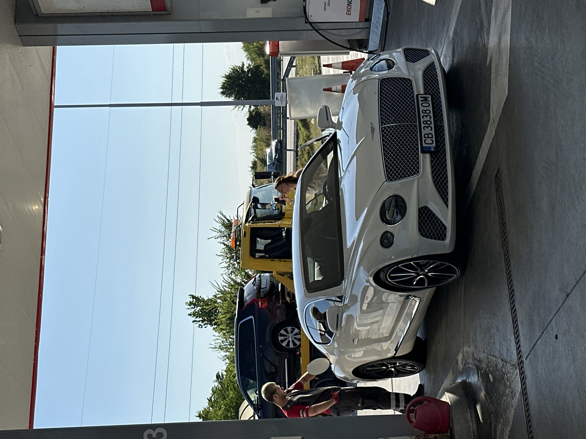 A white Bentley Continental GT is parked at a gas station with its driver's side door open. A person wearing a red and grey uniform, possibly a gas station attendant, is standing next to the car, holding a white disc. A woman is in the driver's seat of the Bentley, looking at her phone. In the background, a yellow tow truck is loaded with a dark SUV. The scene appears to be in Polsko Padarevo, Bulgaria, as suggested by the license plate "CB 3838 OM" which is consistent with Bulgarian plates, and the general environment of a highway-adjacent gas station. The sky is clear and blue, indicating daytime.