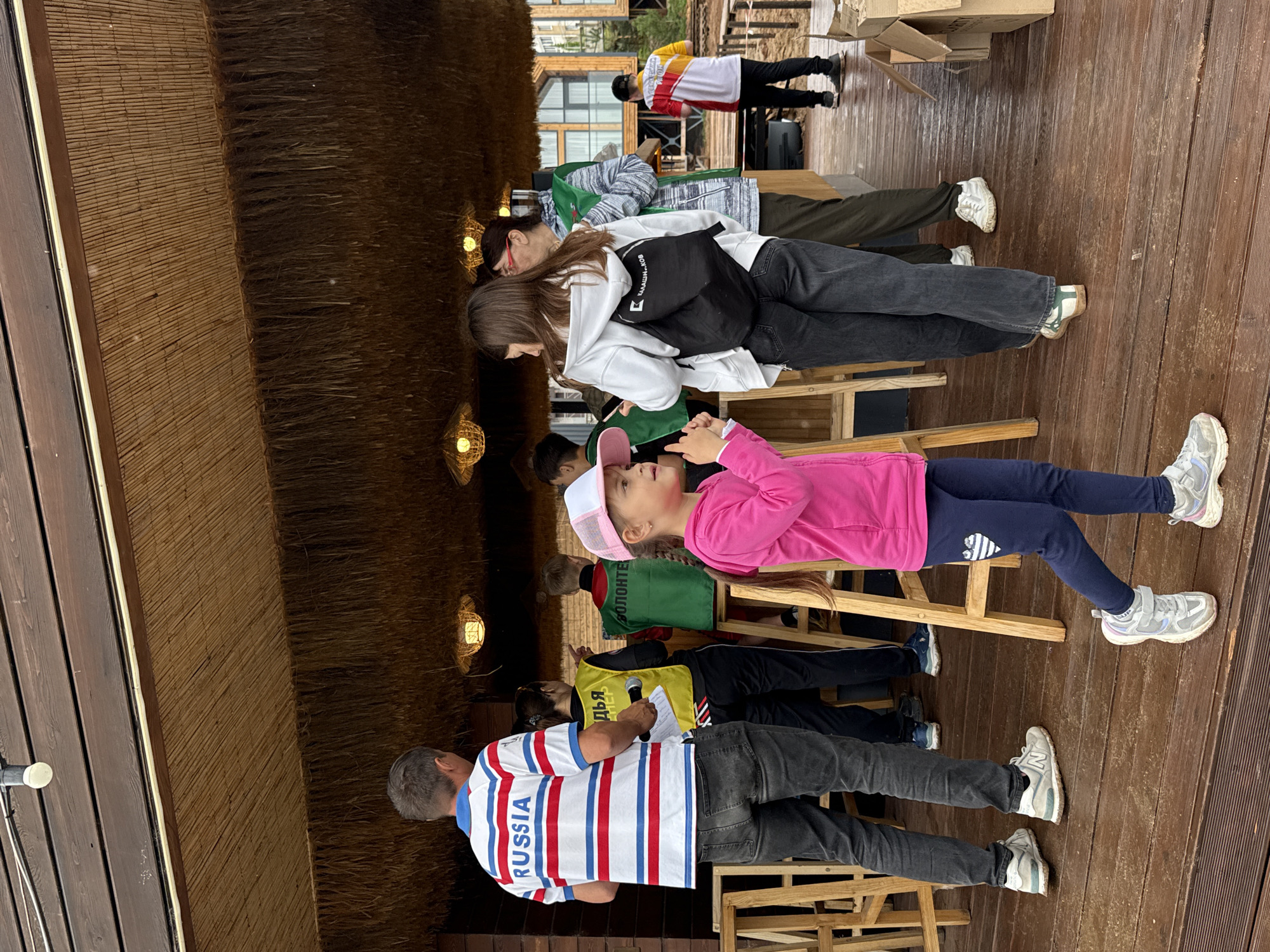 In Votkinsk, Russia, a group of people are gathered under a thatched-roof structure. A man in a striped "RUSSIA" shirt stands on the left, holding a microphone. A young girl in a pink hoodie and baseball cap is in the foreground, looking to the right. Behind them, several individuals in vests, one green with "ВОЛОНТЕР" (Volunteer) visible, are interacting. Another person is wearing a black backpack with Cyrillic lettering. The setting appears to be an outdoor event, possibly a festival or gathering, with wooden furniture and rustic architectural elements. The ground is made of wooden planks. In the background, modern residential buildings are visible.