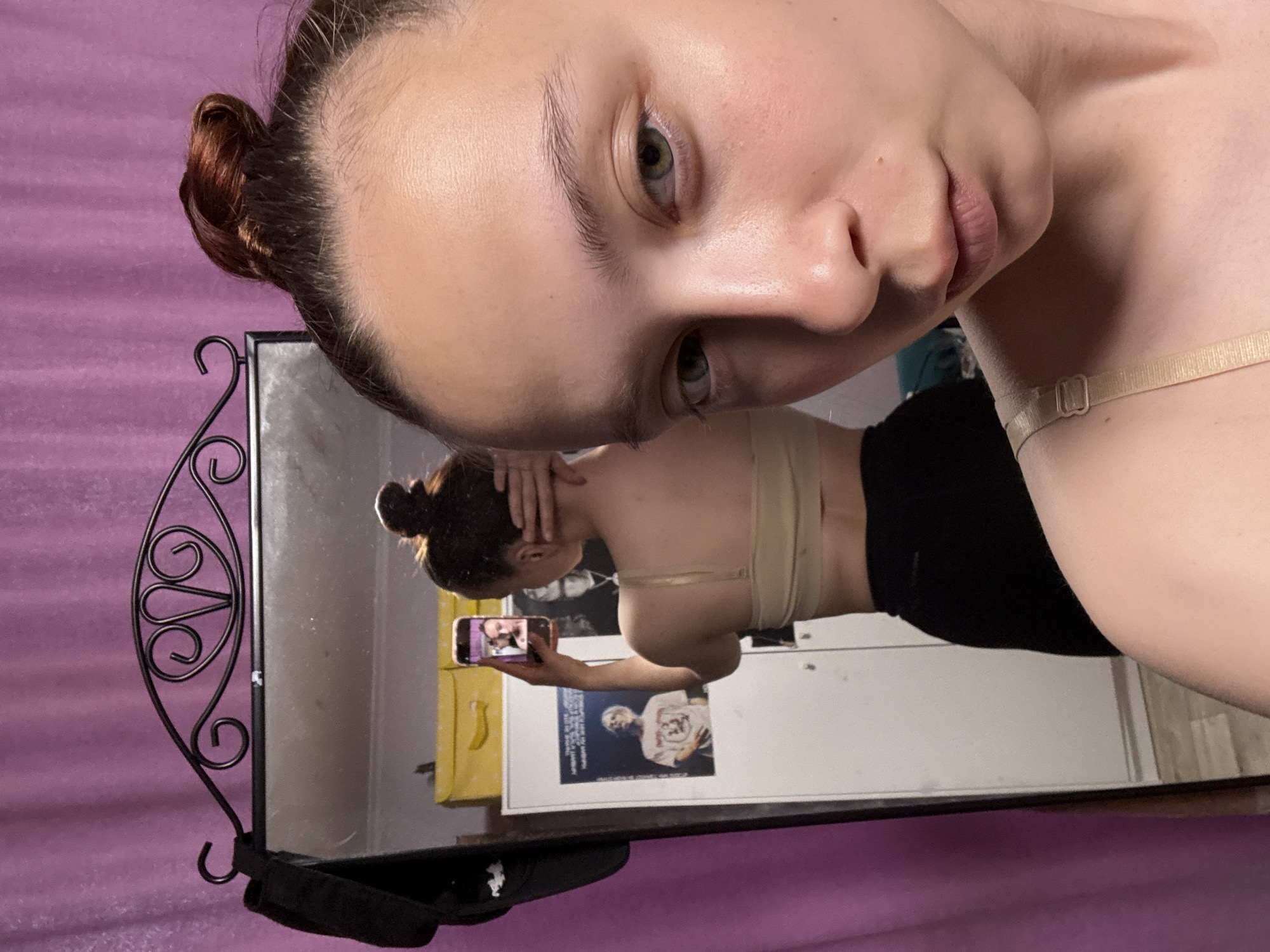 A young woman with fair skin and brown hair in a bun takes a selfie in a mirror. The reflection in the mirror shows the woman holding a phone and touching her hair, wearing a beige top and black shorts. Behind her is a white door with posters. The wall behind the mirror is purple. The mirror has a black, ornate frame. There is no evidence of a specific location in the city of Прикамский, Russia.