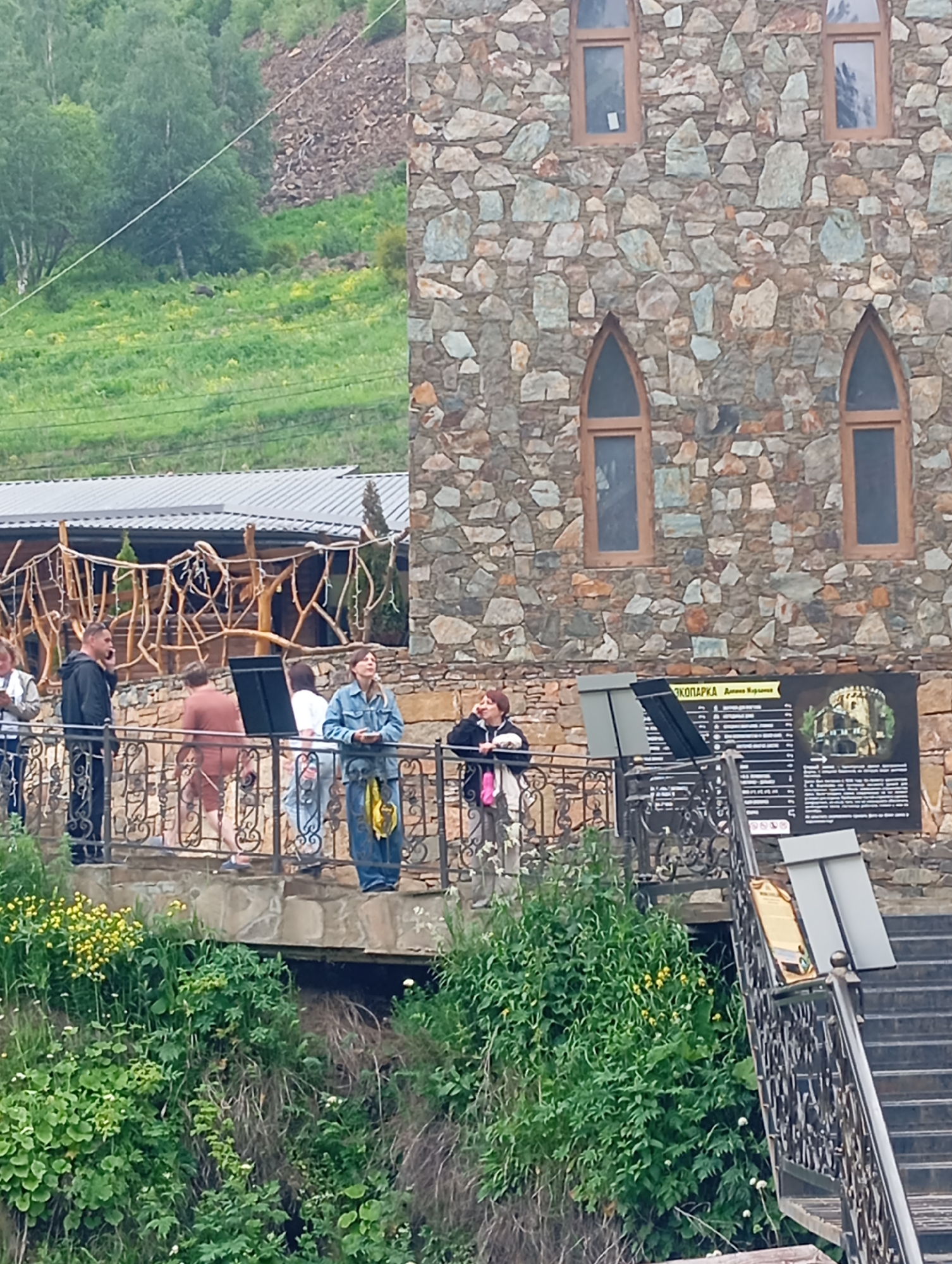 The image depicts an outdoor scene with a stone building and a group of people. The building has a rustic facade made of irregularly shaped stones and features arched windows with wooden frames. In the foreground, a group of individuals is standing on a balcony or walkway overlooking a lush, green hillside. Some of the people are engaged in conversation, while others are looking around. A sign with text and an illustration is attached to a railing. The environment is natural, with trees and vegetation visible on the slopes. The overall impression is of a tourist attraction or a scenic spot.