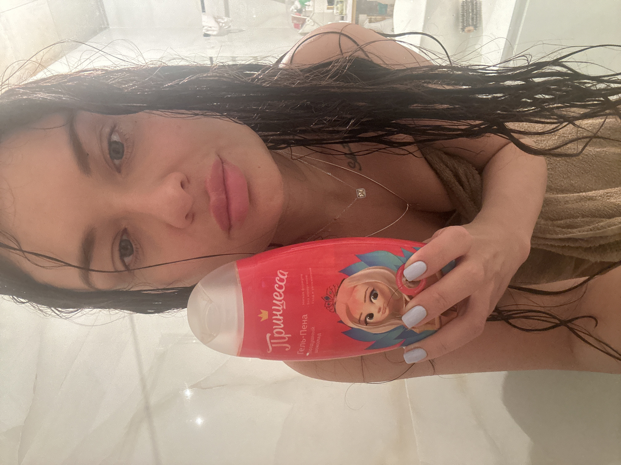 A close-up, tilted portrait shows a woman in what appears to be a bathroom or shower setting. Her dark, long, wet hair is splayed around her, indicating she has recently showered. She is wrapped in a light brown towel, visible on her shoulder and chest. Her face is turned towards the camera, with a soft expression, full lips, and dark eyes. She wears a delicate silver necklace with a square pendant and has light-colored nail polish on her fingers.
She holds a red and pink bottle of bath product in her right hand, positioned prominently in the foreground. The bottle features a cartoon illustration of a blonde princess with a crown. Visible text on the bottle is in Russian and reads: "Принцесса" (Princess), "Гель-пена" (Gel-foam), and "Облачный шоколад" (Cloudy chocolate), likely referring to its scent. Smaller text includes "Мягкая формула" (Soft formula), "без аллергенов" (hypoallergenic), and "уход и увлажнение" (care and hydration). The background consists of light-colored, possibly tiled, surfaces, characteristic of a bathroom.