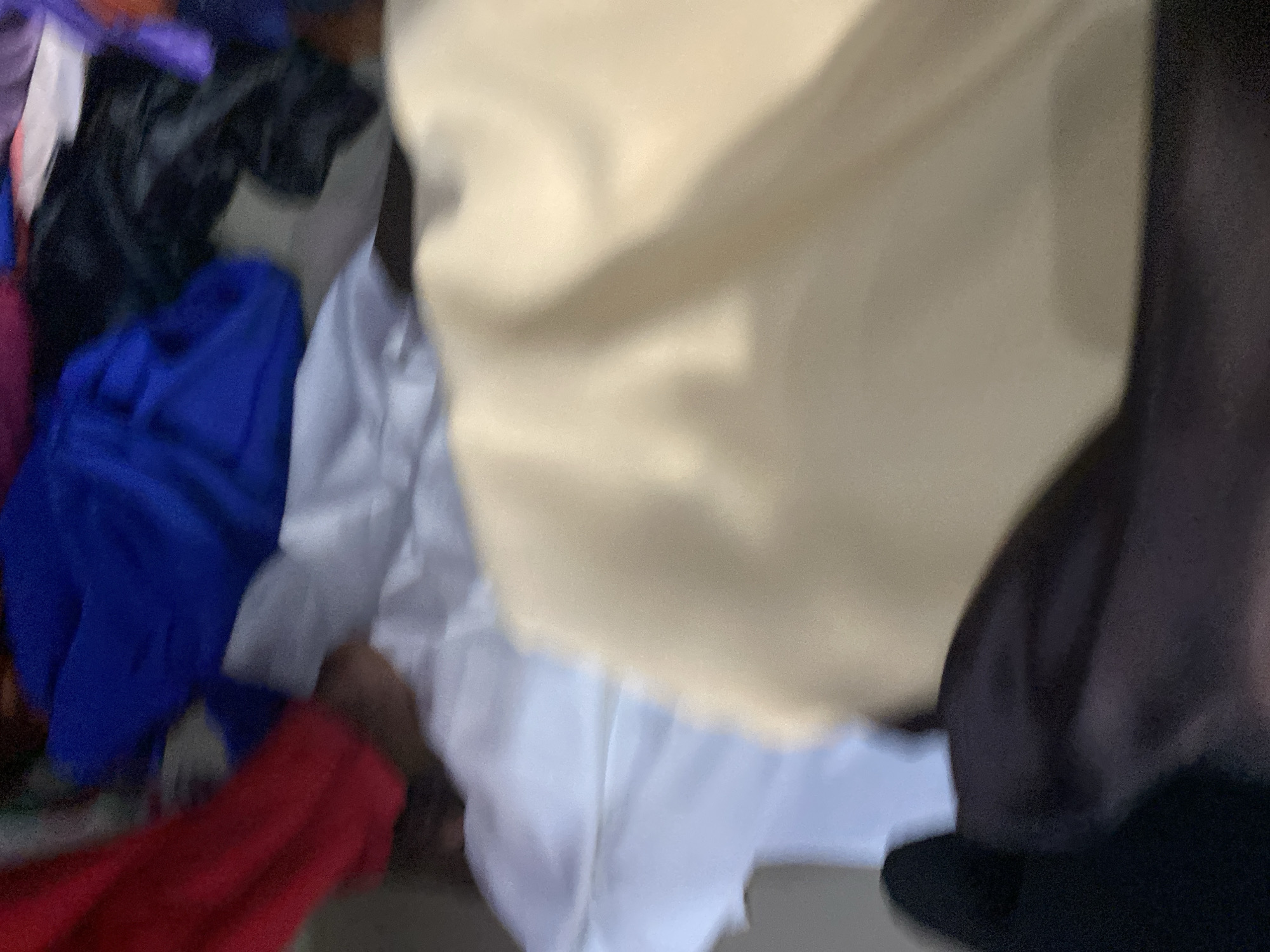The image shows a pile of fabric, with a prominent beige-colored piece in the foreground, draped in soft folds. Behind it, white fabric is visible, followed by a bright blue fabric and a red fabric. Towards the right, dark colored fabric, possibly black or dark purple, is also present. The lighting is soft, creating subtle shadows and highlights on the fabric textures. The scene appears to be indoors, with the fabrics likely stacked or arranged in a storage or display area. There is no discernible urban environment or specific location in Jalingo, Nigeria visible in the image.