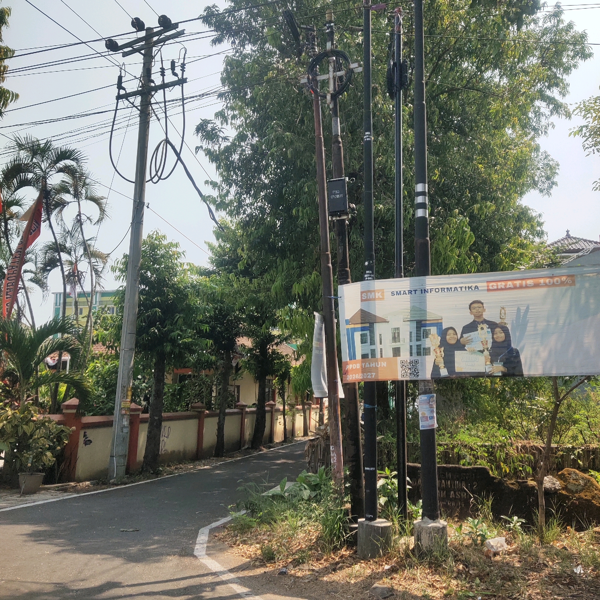 The image shows a street scene in Sragen, Indonesia, with several utility poles lining the side of the road. Palm trees and other lush green foliage are abundant, suggesting a tropical or subtropical climate. A building, possibly a school, is visible in the background, featuring light green and yellow paint. A large banner advertising "SMK SMART INFORMATIKA GRATIS 100%" is prominently displayed on one of the utility poles. The banner includes a picture of three students holding trophies, indicating an educational institution. A red flag with text is also visible on the left side of the image. The road appears to be asphalt, with white lines marking its edge. The overall impression is of a well-maintained street in a residential or semi-commercial area of Sragen.